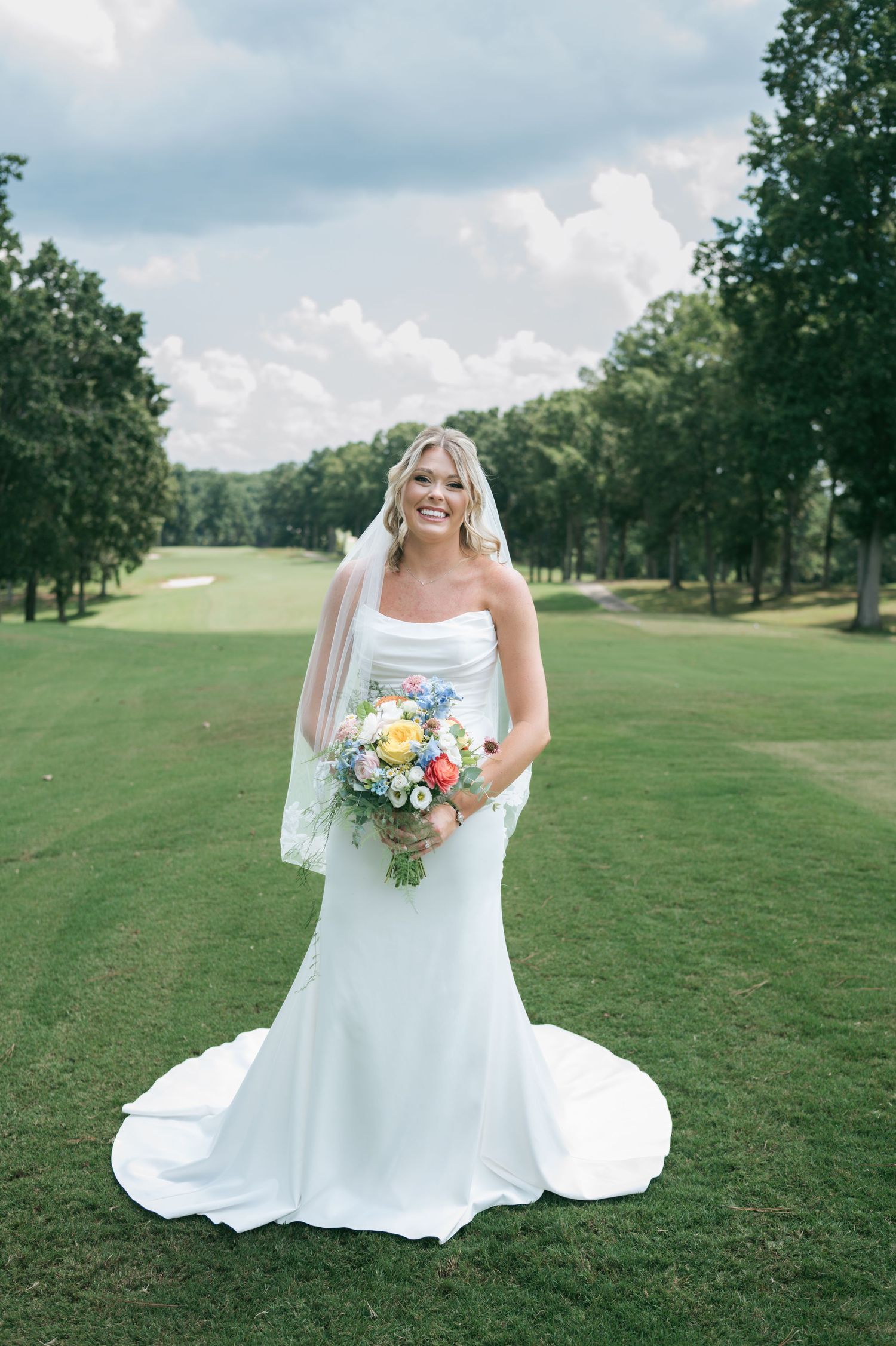 Radiant bride in sleek strapless gown holds colorful bouquet while posing on lush golf course under cloudy sky, wedding photographer NC