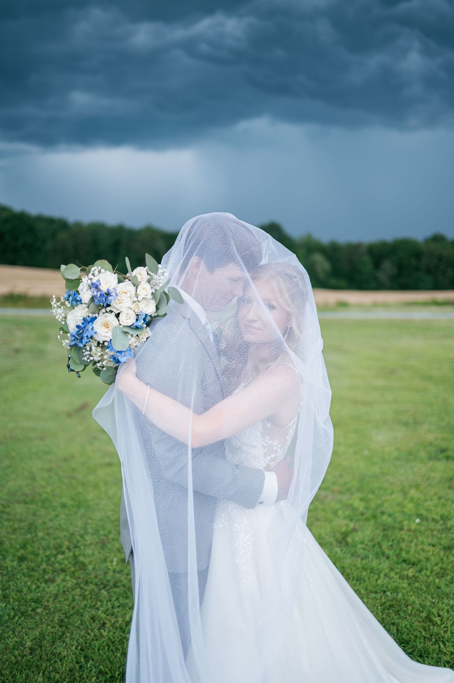 Bride and groom share romantic kiss under flowing veil outdoors with dramatic stormy sky and green field backdrop, wedding photographer NC