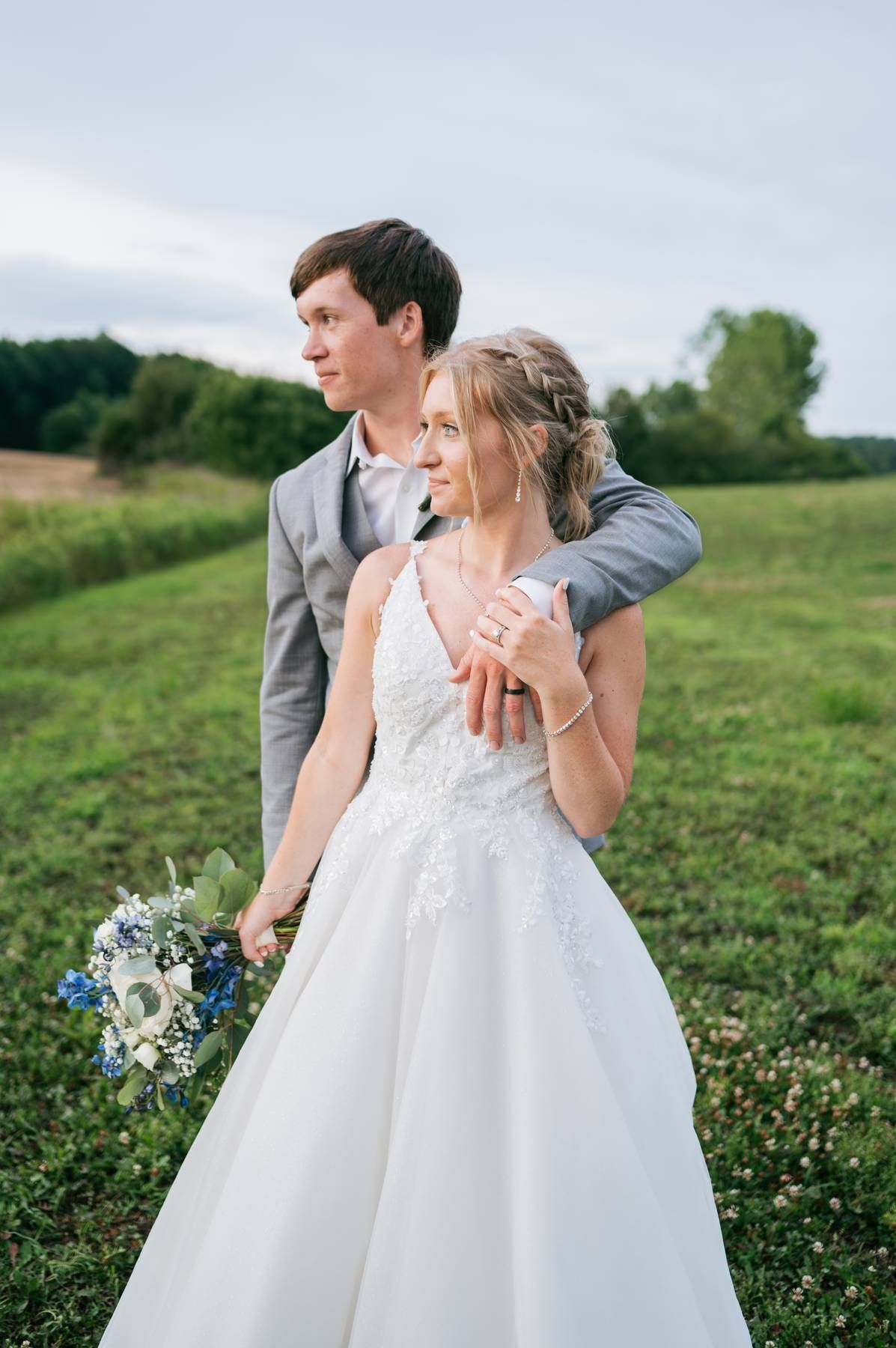 Bride and groom embrace in a lush green field, she holds blue and white bouquet at sunset. Asheboro NC