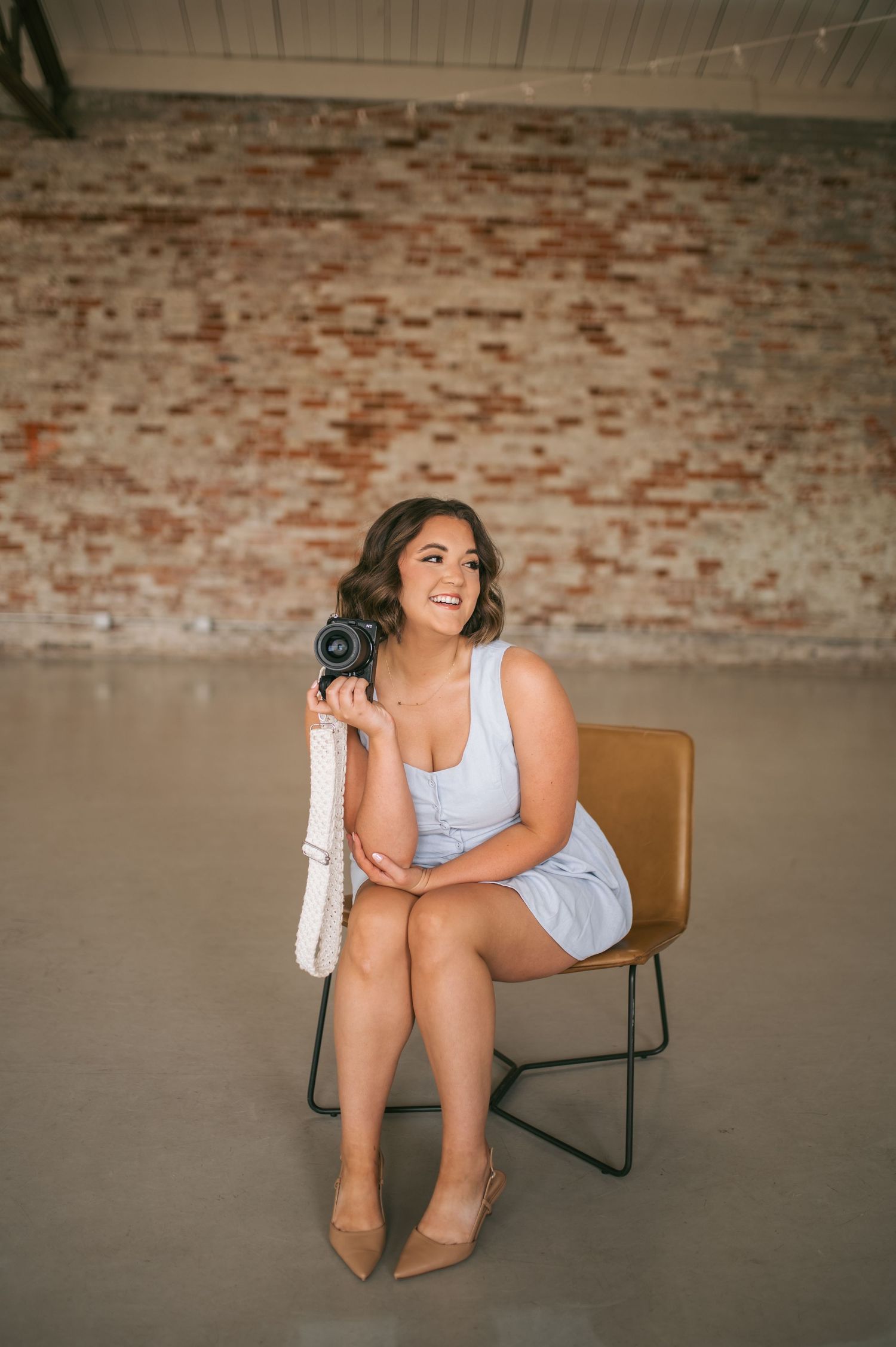 Smiling brunette photographer in white dress sits on chair holding camera in front of rustic brick wall, portrait photographer NC