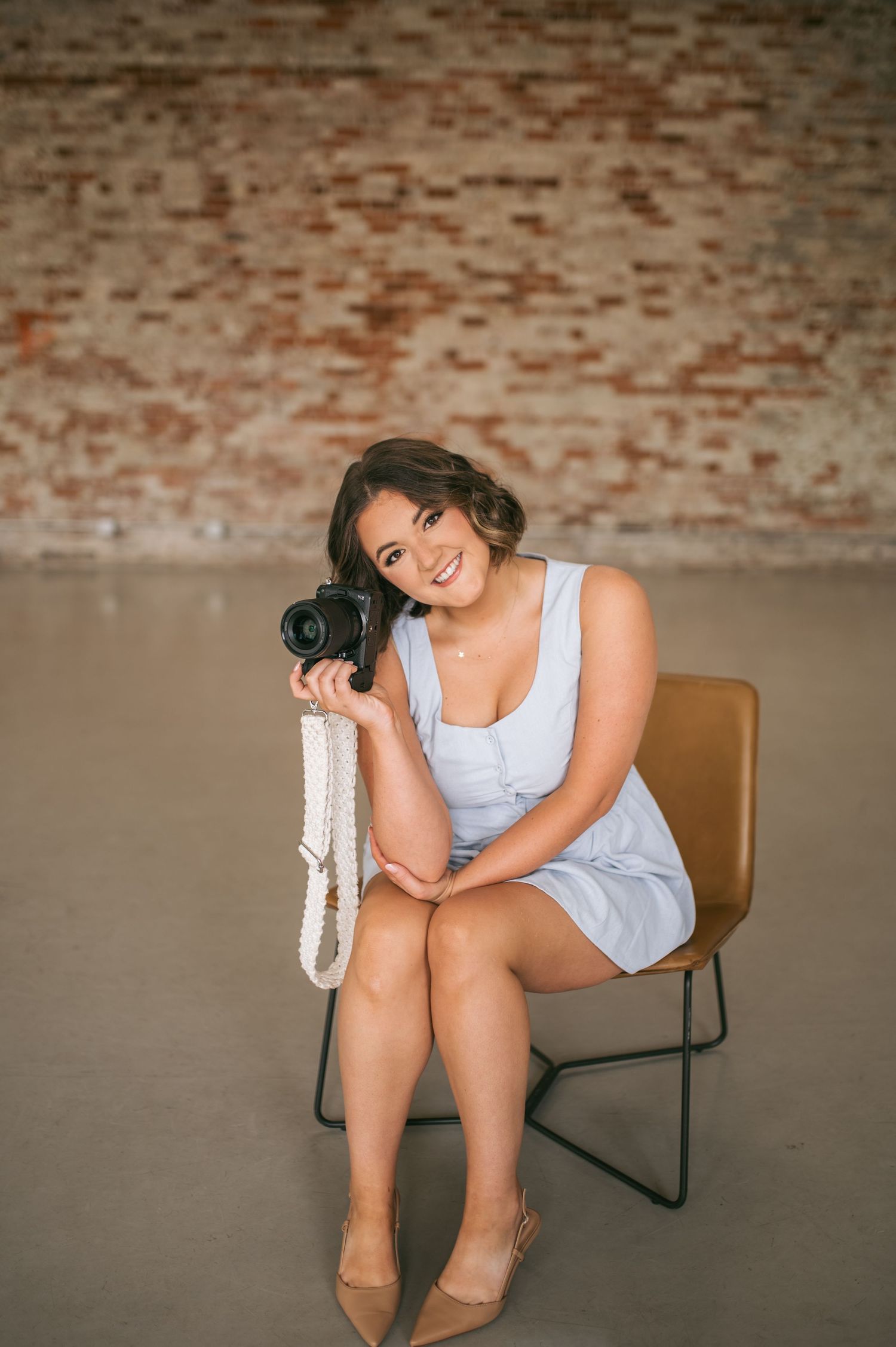 Smiling young woman in blue dress sits on chair holding camera with brick wall background, portrait photographer NC