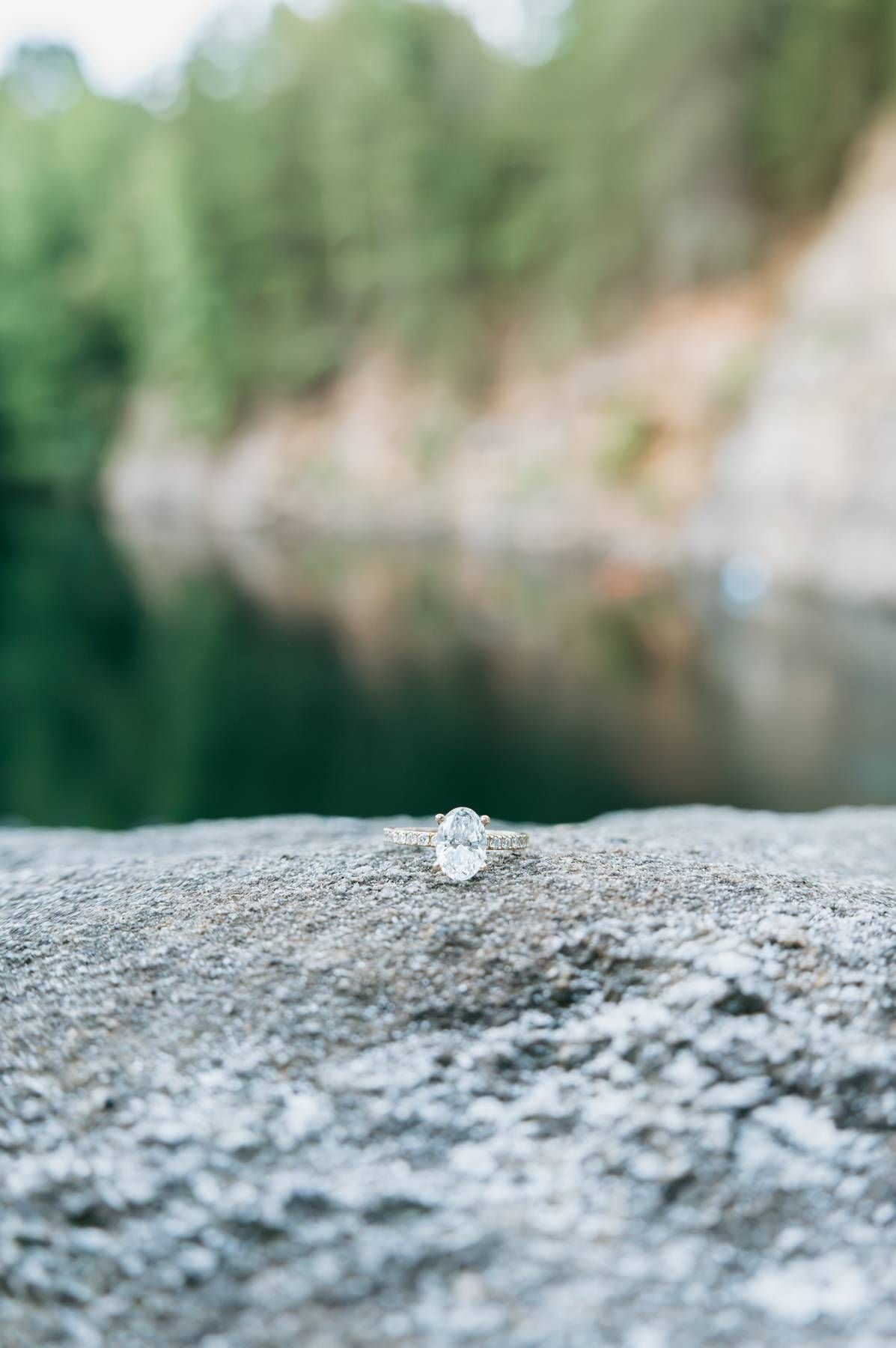 Oval diamond solitaire engagement ring displayed on rocky surface with blurred lake and trees background in North Carolina