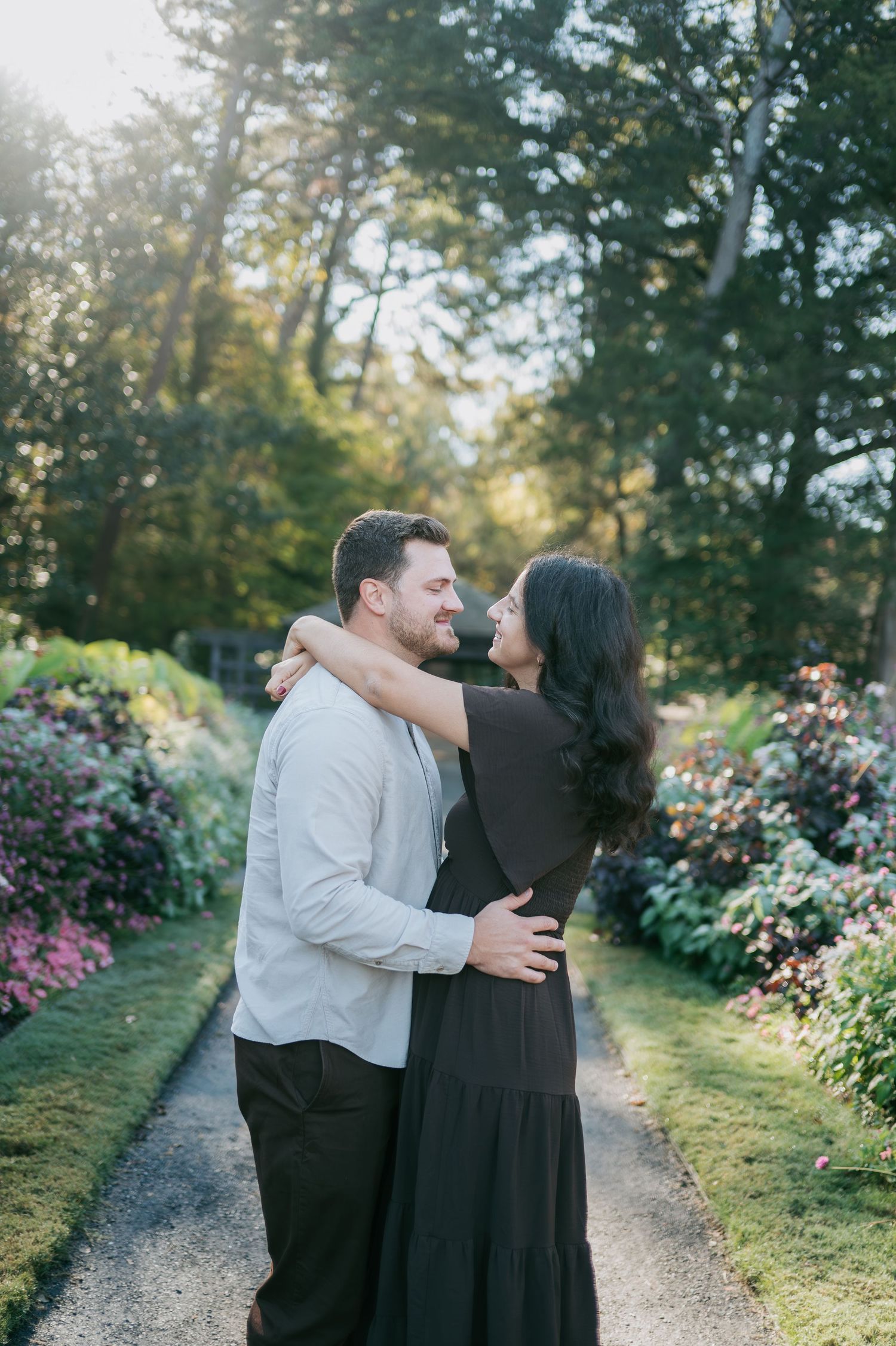 Couple embracing and smiling at each other along a flower-lined garden path bathed in warm golden sunlight, engagement photographer NC
