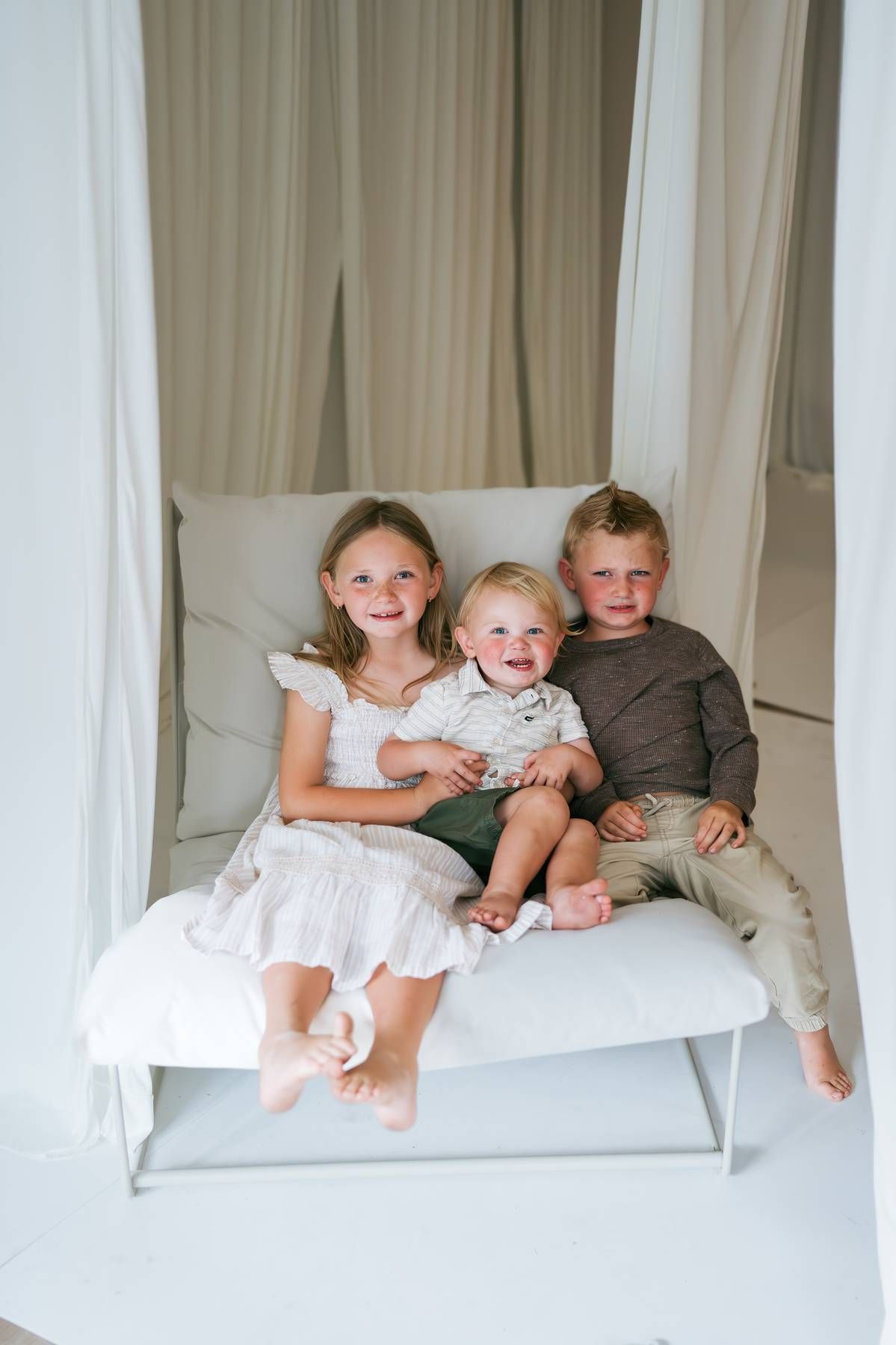 Three young siblings sitting together on a white bench smiling for a portrait, family photographer Asheboro NC