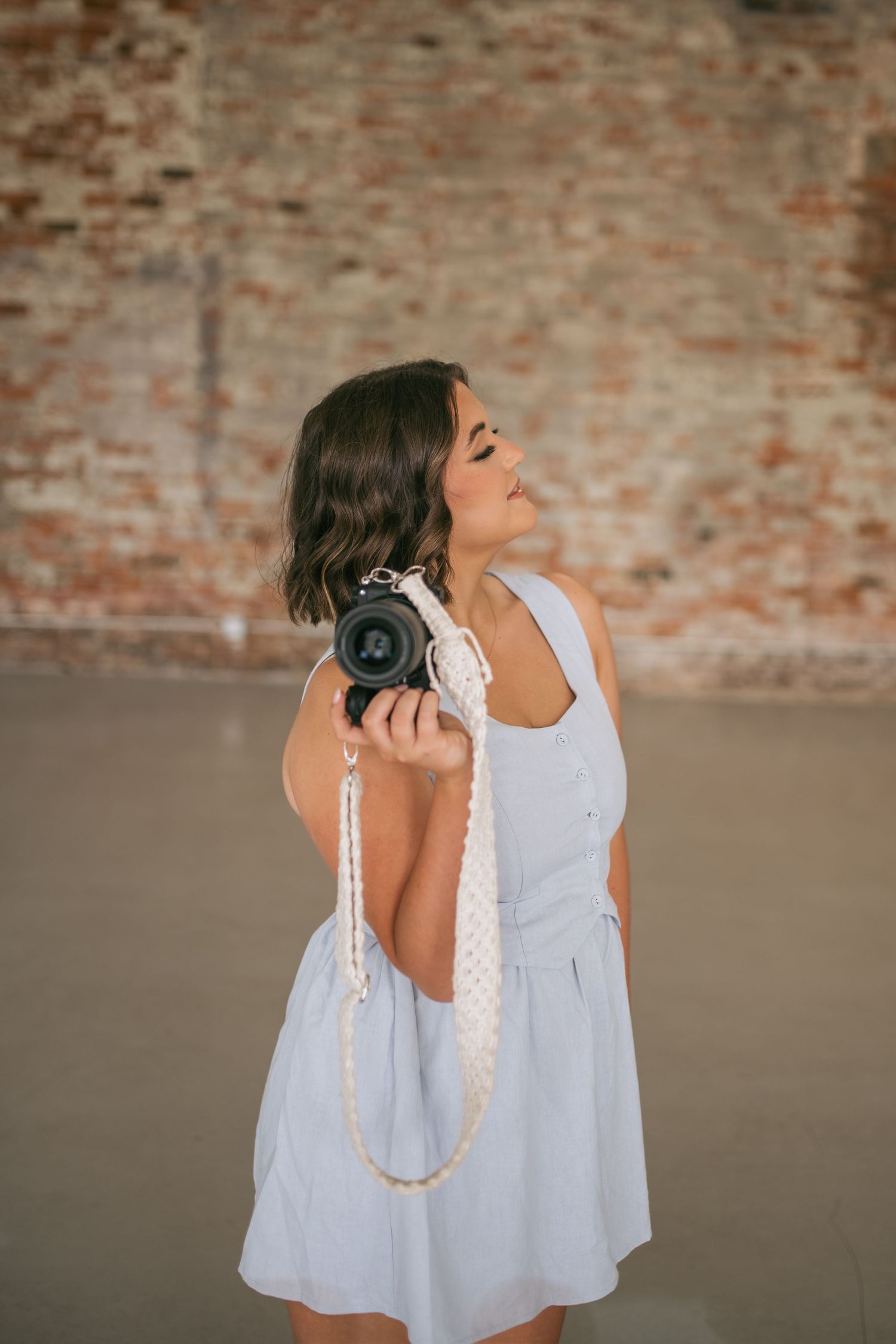 Brunette female photographer in light blue dress poses with camera against rustic exposed brick wall backdrop, wedding photographer NC