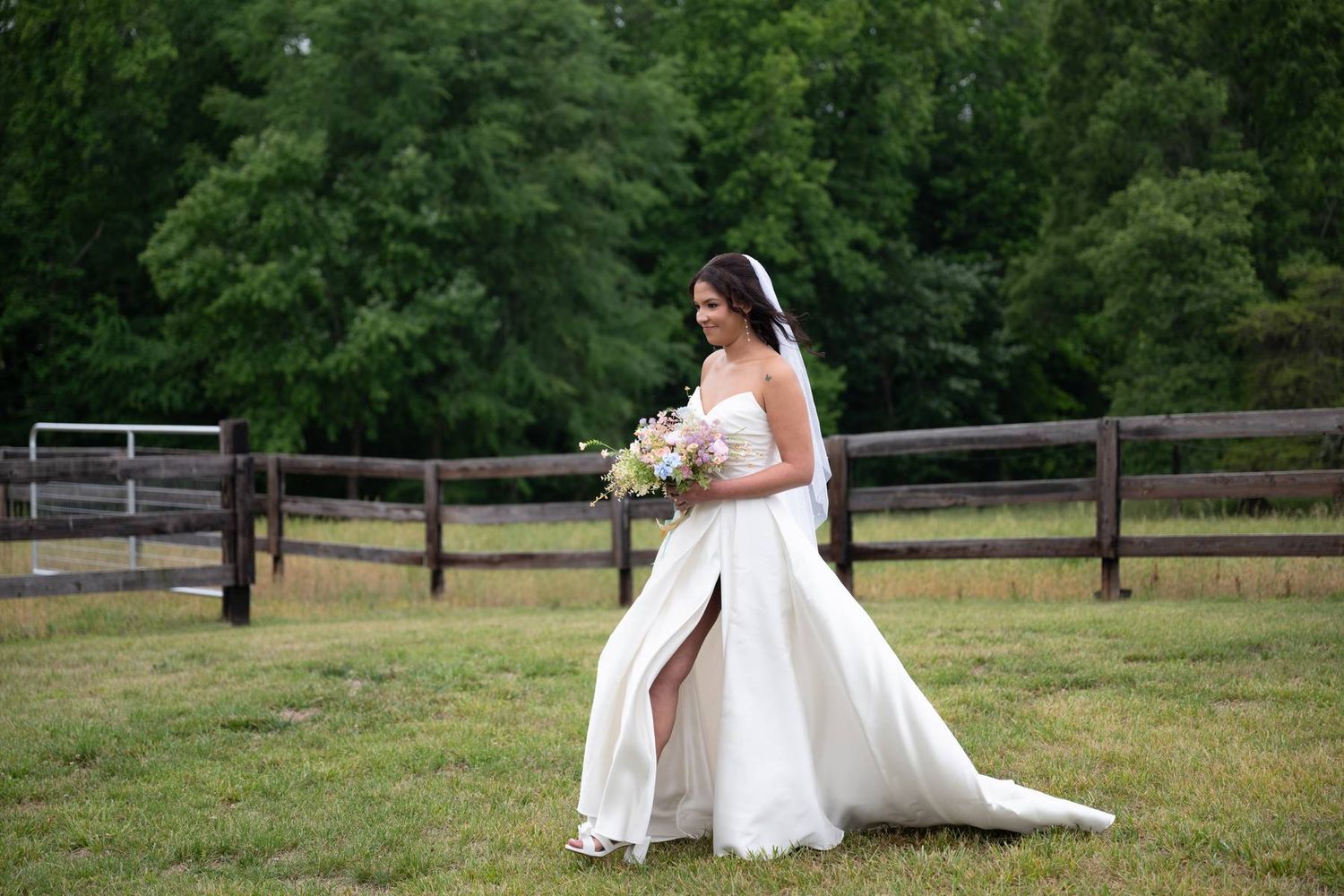 Bride in white gown with slit holding floral bouquet walking near rustic wooden fence in North Carolina