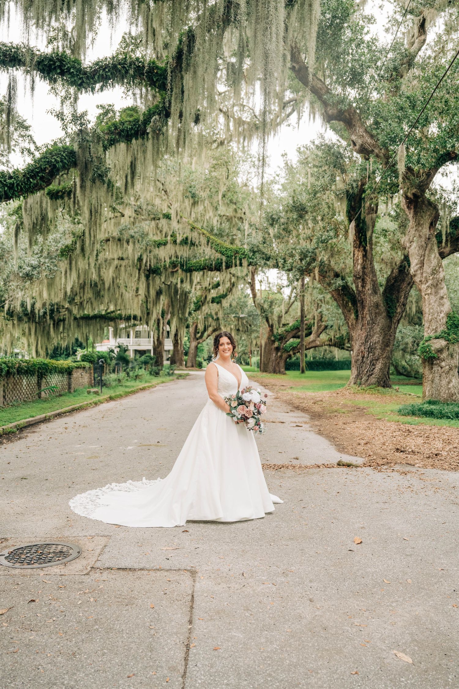 A bride in a flowing white gown poses on a scenic path lined with Spanish moss-draped oak trees in a Southern garden.