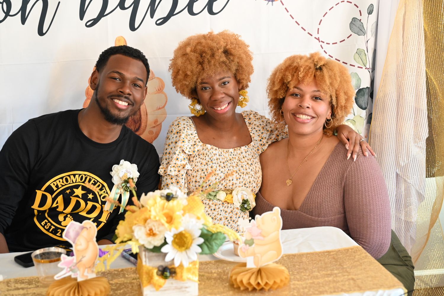 Three people smile together at a bee-themed baby shower with sunflower decorations and an oh babee backdrop.