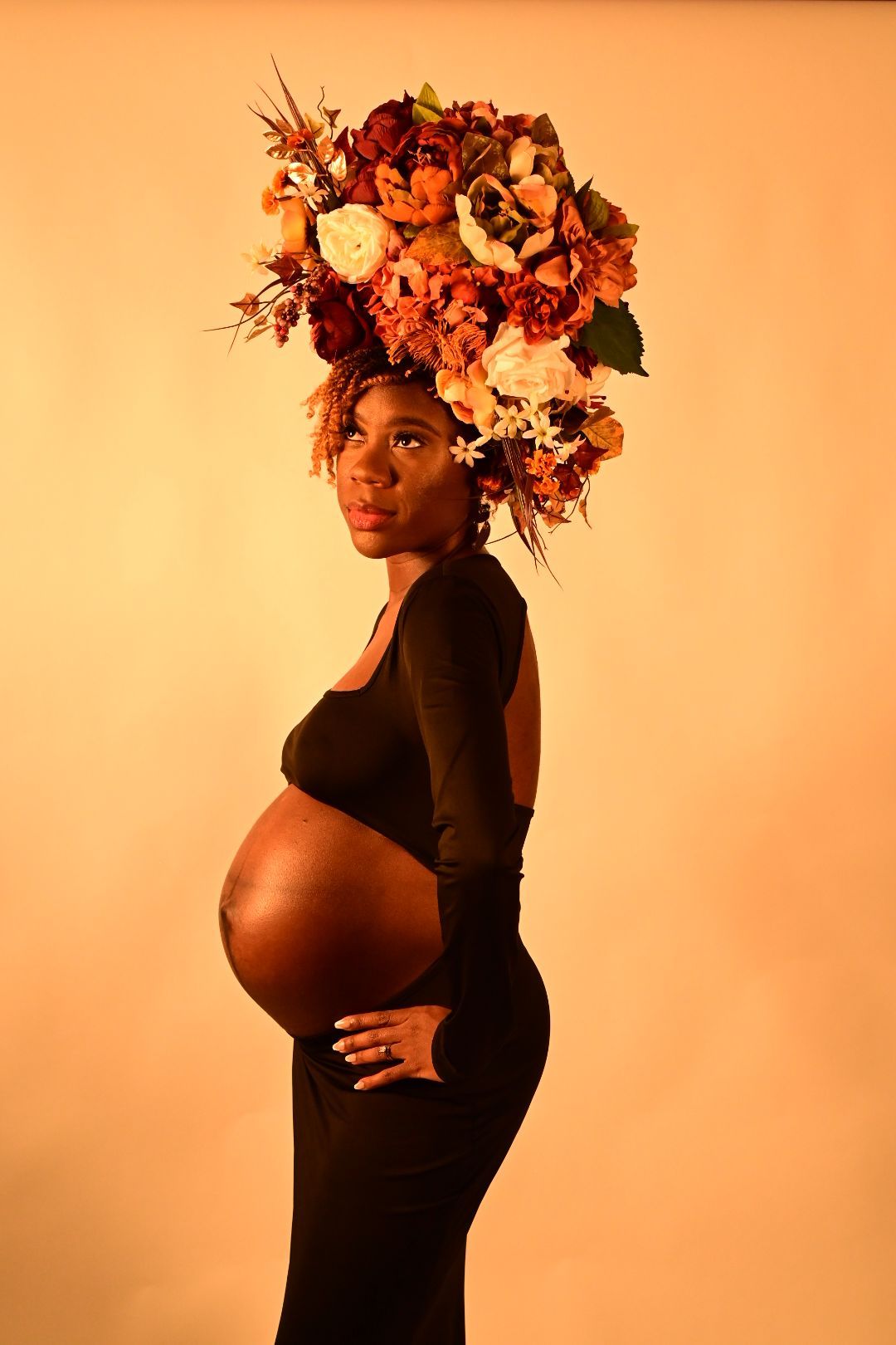 Pregnant Black woman in black dress wearing large floral crown, posing sideways showing baby bump.