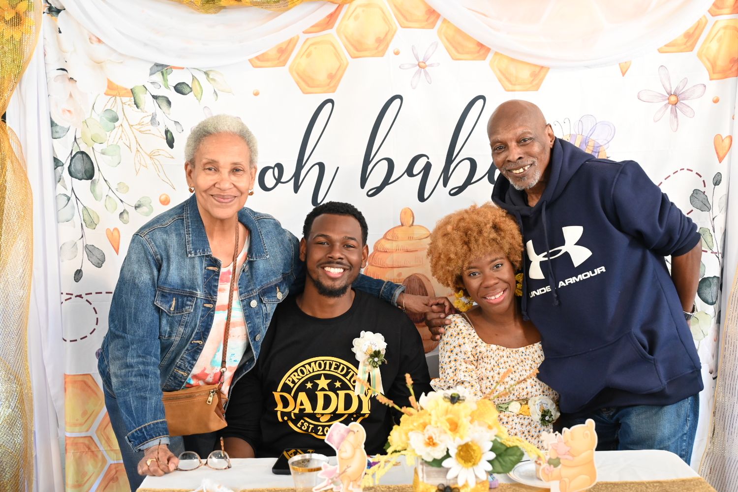 Four smiling people gathered at a honeybee themed baby shower with an oh babee backdrop.