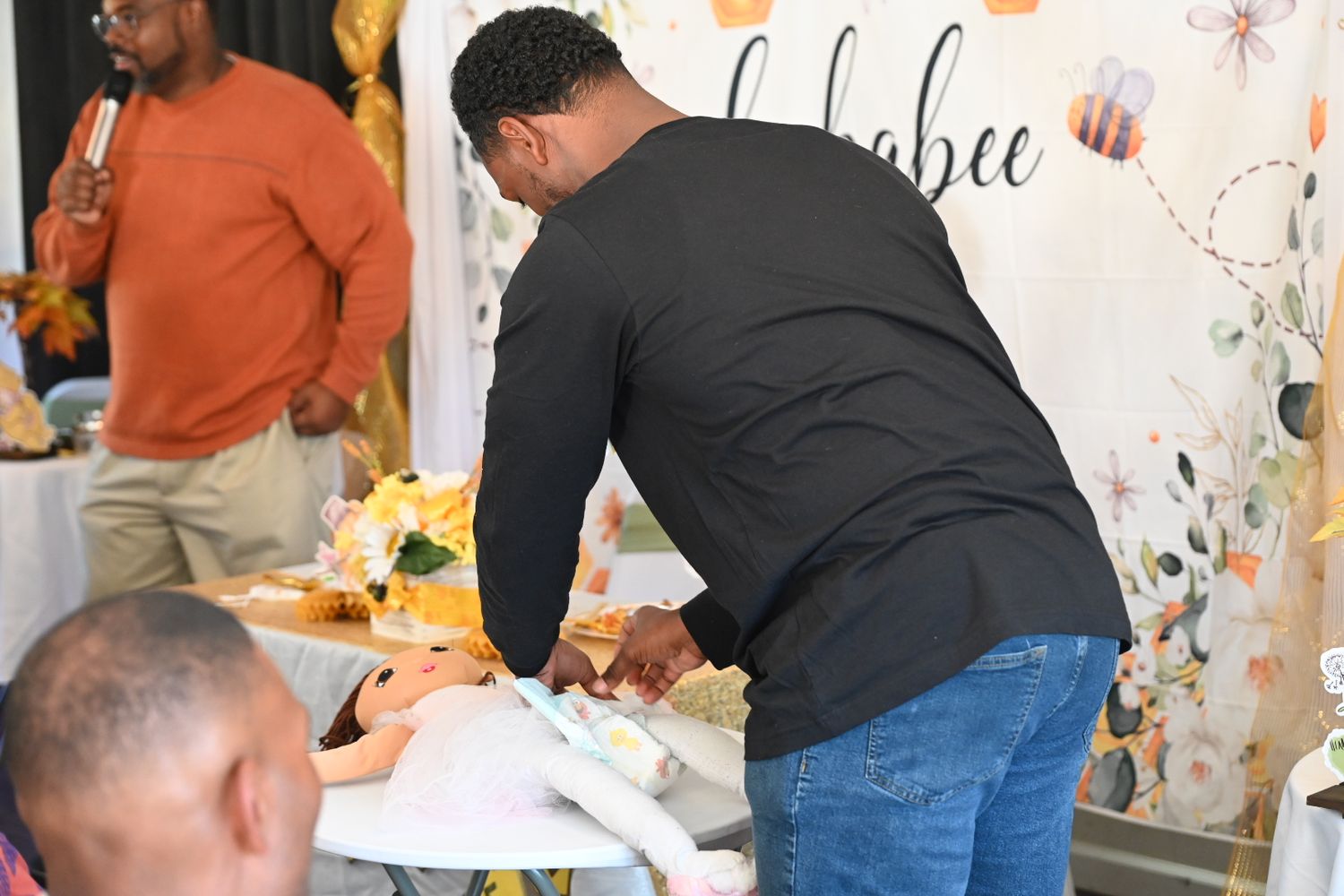A man in black shirt leans over a baby shower gift table in front of a floral bee-themed backdrop.