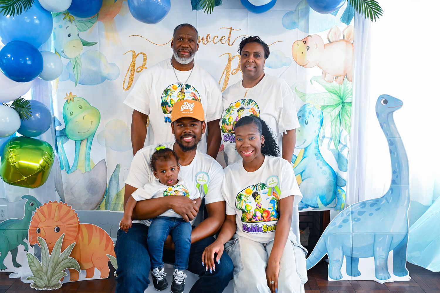 Family posing at a dinosaur-themed baby boy birthday party with blue balloon arch backdrop.
