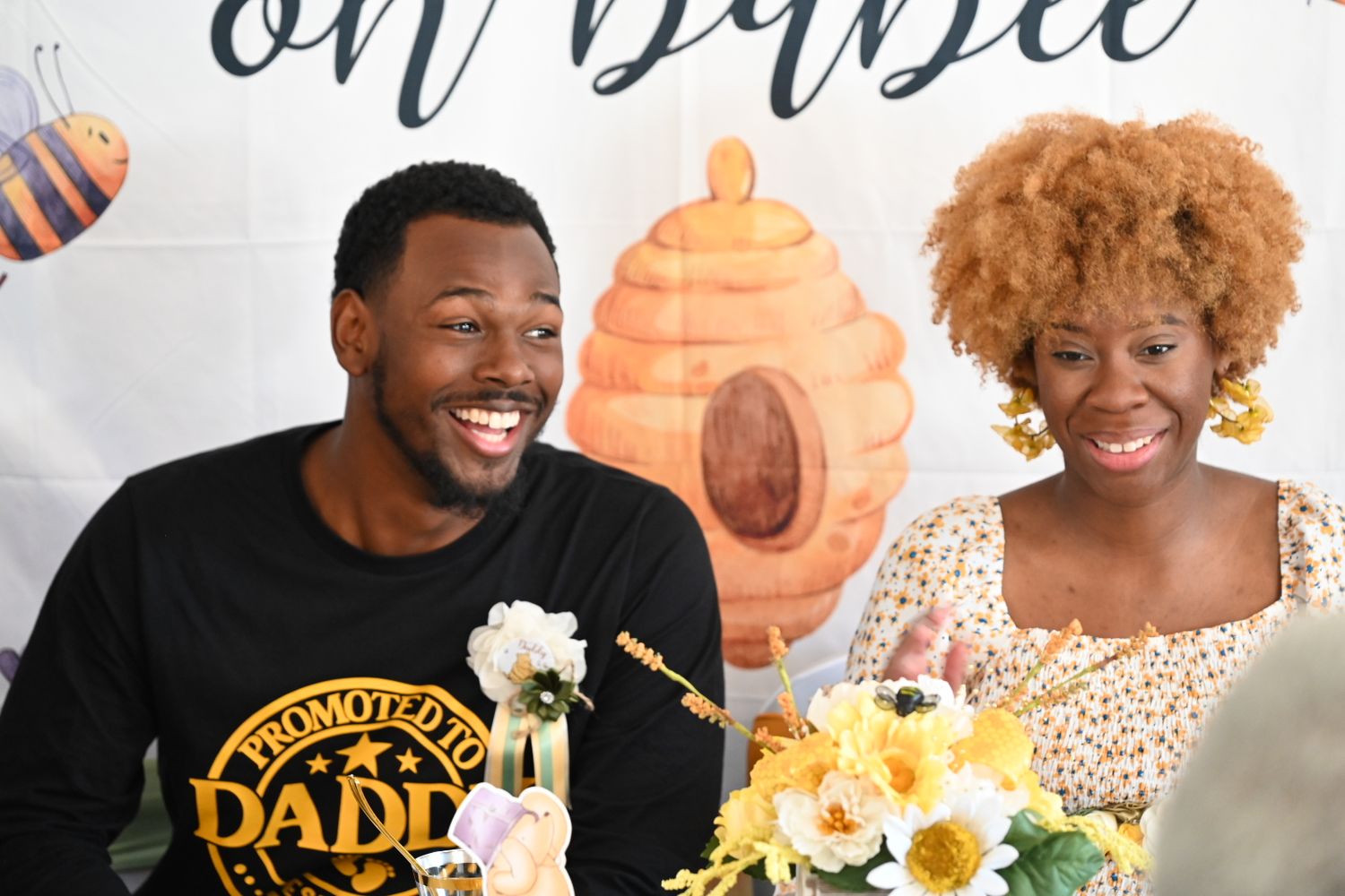 A man wearing a Promoted to Daddy shirt laughs with a woman at a bee-themed baby shower.