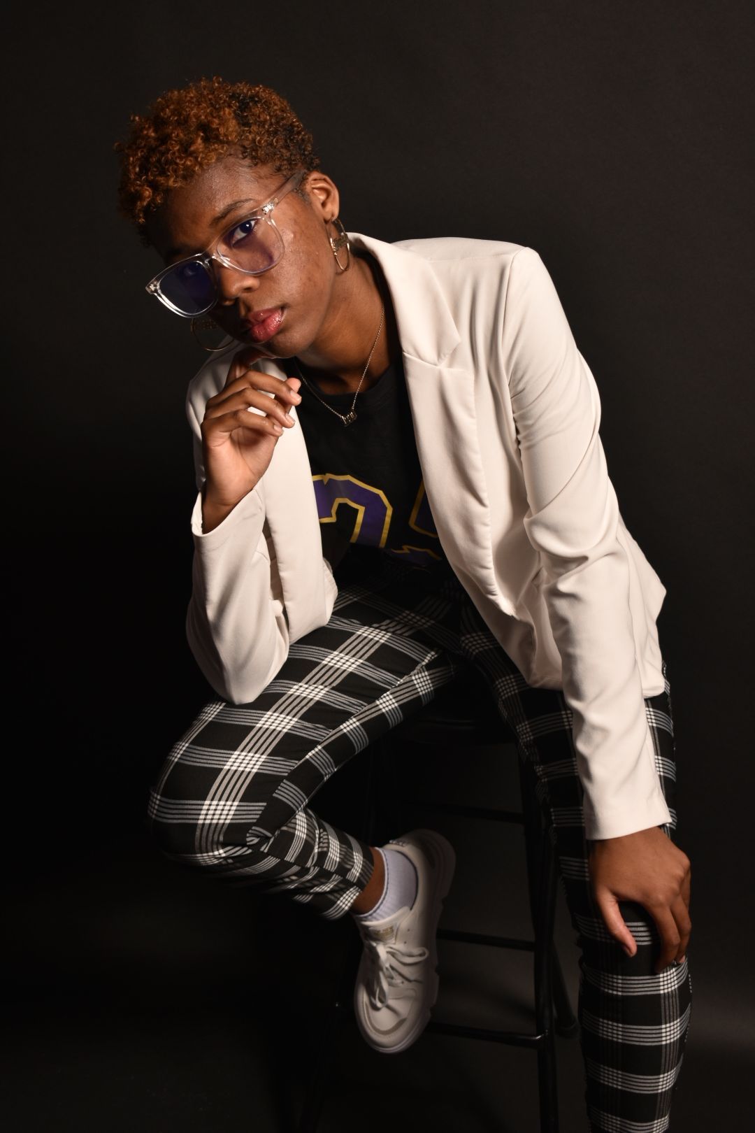 Fashionable woman in white blazer and plaid pants sits on stool adjusting clear glasses in studio.