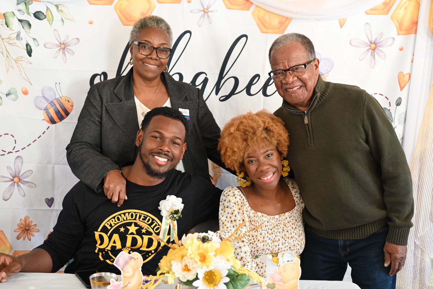 Four adults posing together at a baby shower, man wearing Promoted to Daddy shirt seated at table.