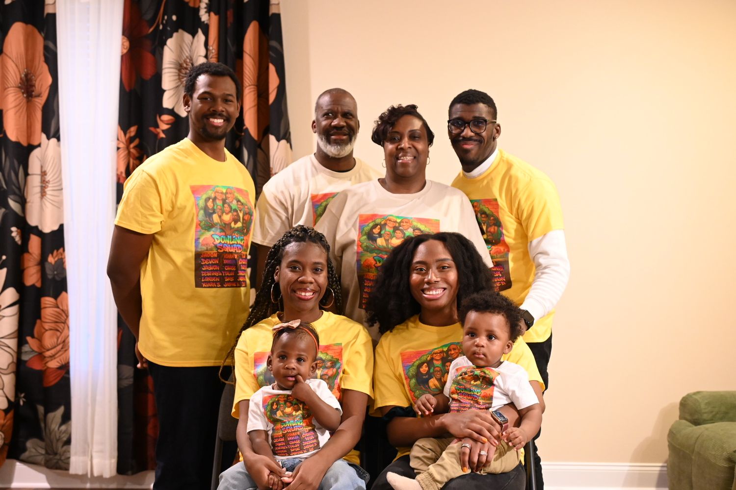 Family group photo with adults and babies wearing matching yellow graphic t-shirts, posing together indoors.