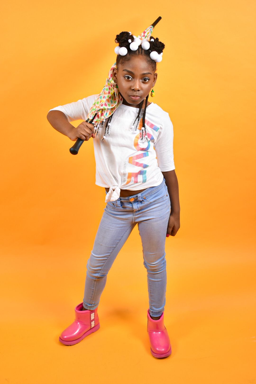 Young Black girl with colorful braids holding baseball bat, wearing jeans and pink boots on orange background.