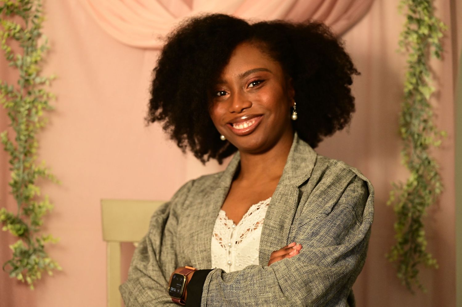 Smiling Black woman in gray blazer poses with arms crossed against pink floral backdrop.