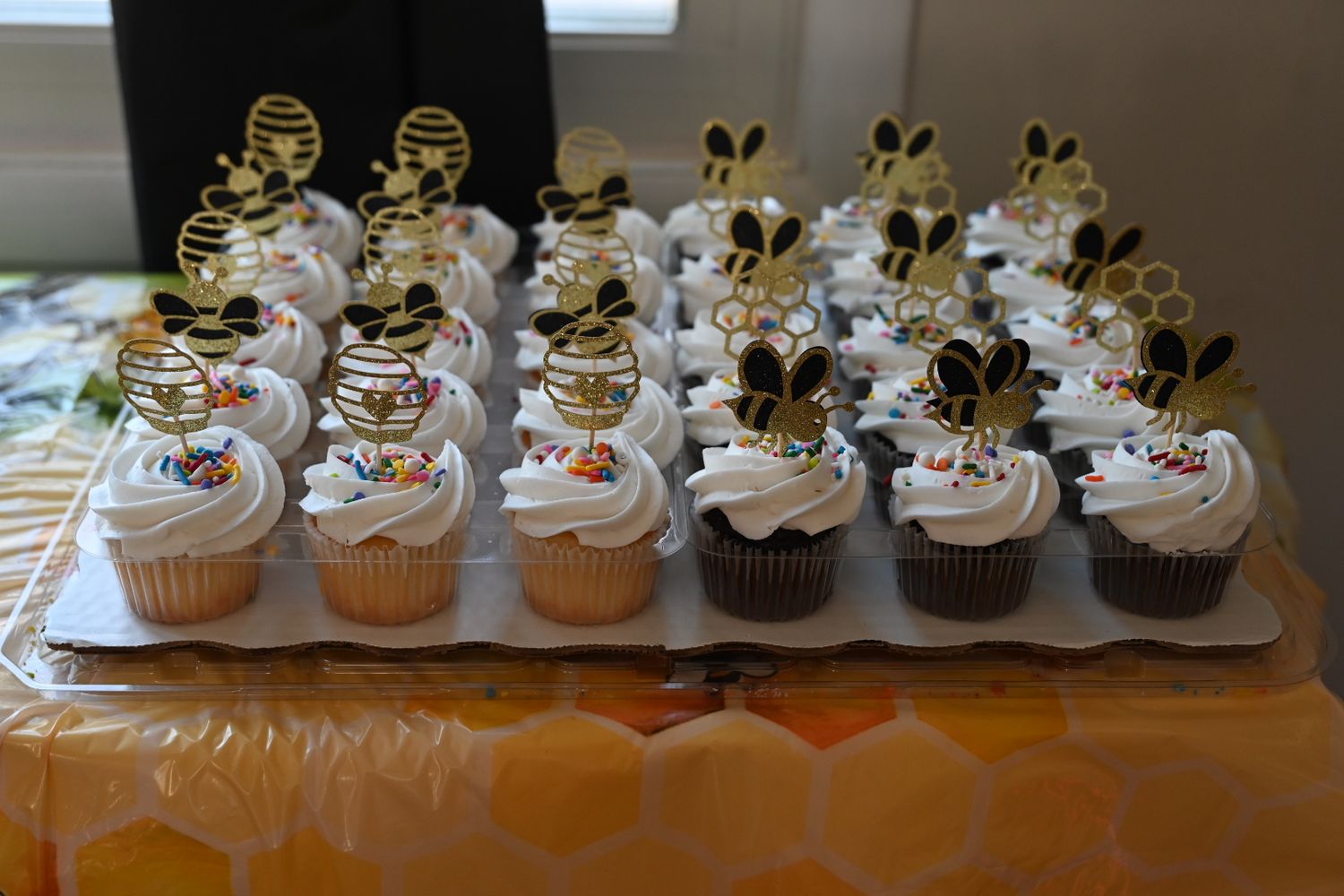 Bee-themed baby shower cupcakes with white frosting and black and gold bee toppers on a tray.
