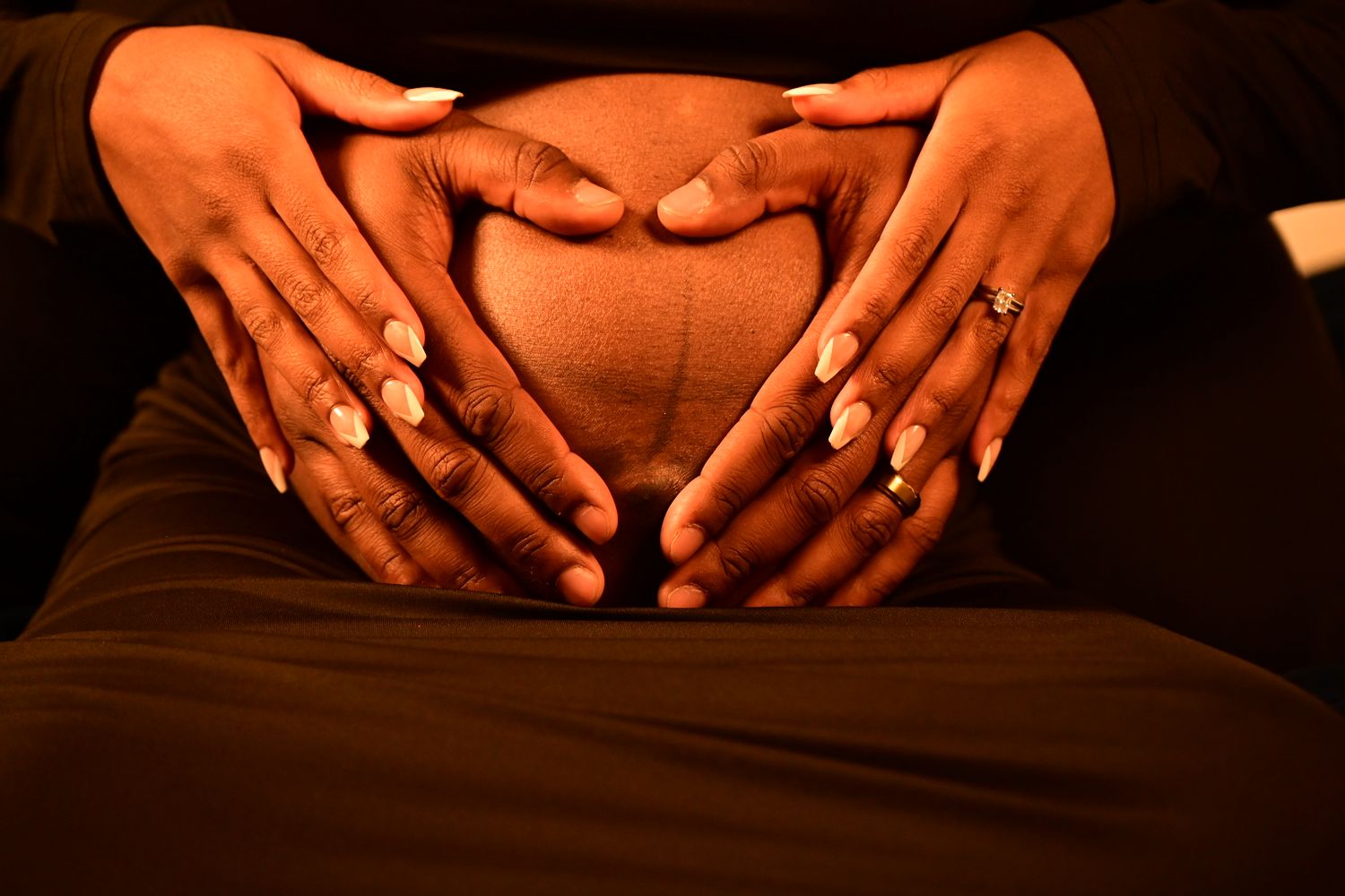Two pairs of hands forming a heart shape on a pregnant belly in warm lighting.