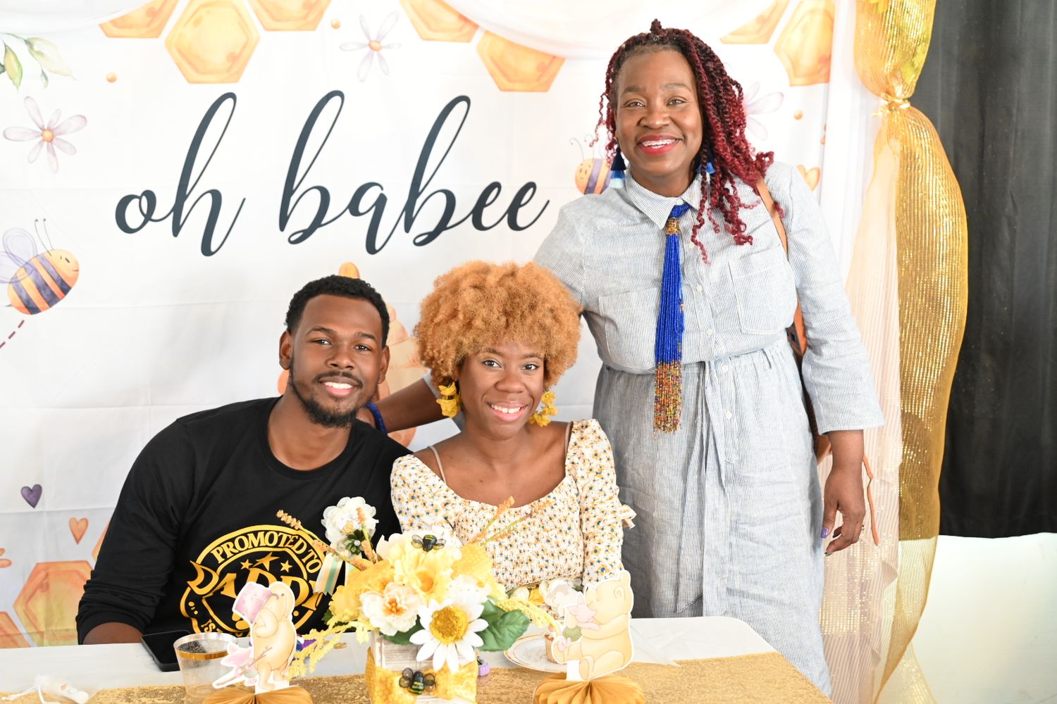 Three people smiling at an oh babee baby shower with yellow floral arrangements on the table.