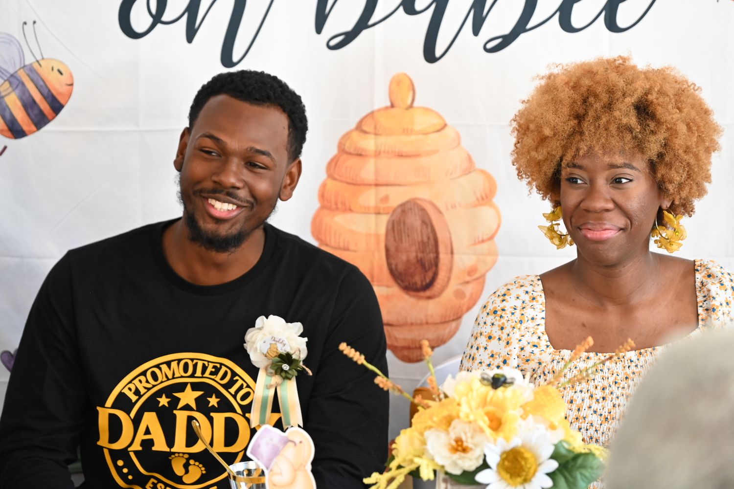 A man in a Promoted to Daddy shirt smiles with a woman at a bee-themed baby shower table.