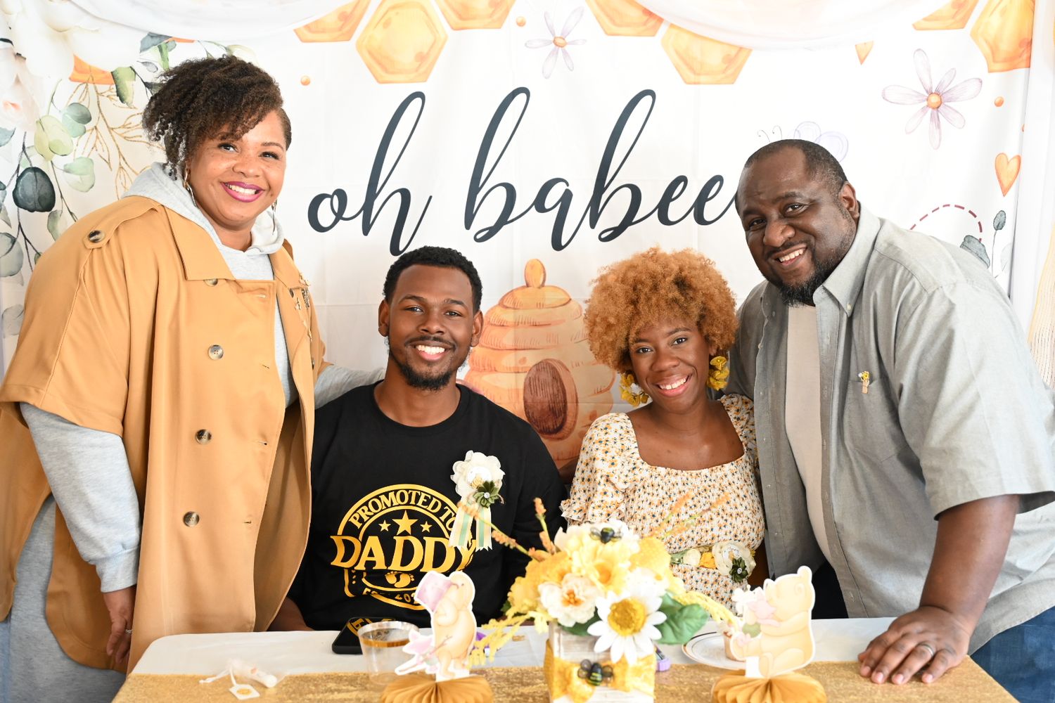 Four people pose together at a bee-themed baby shower with honeybee decorations and oh babee banner.