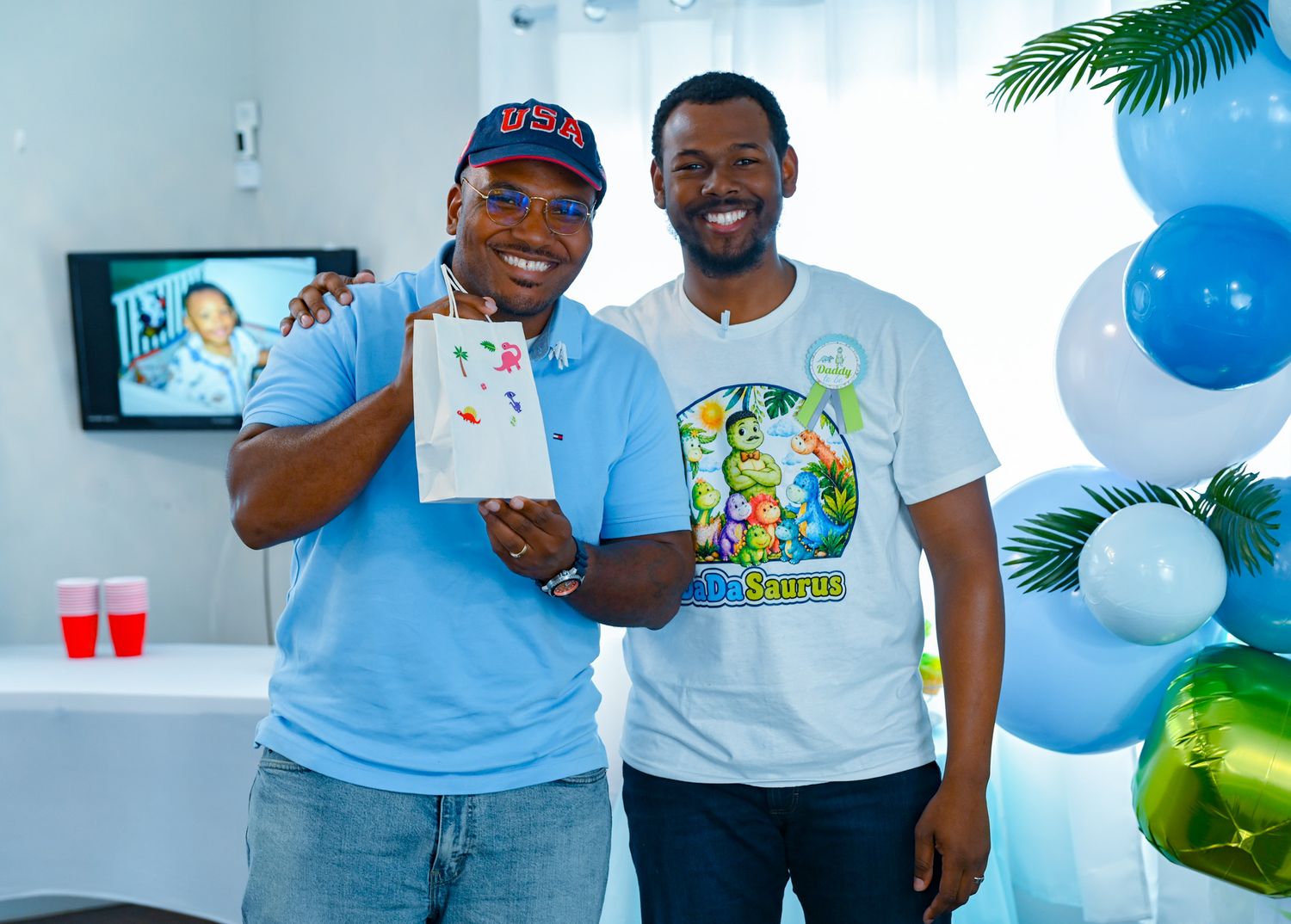 Two Black men smile together holding a card at a baby shower with blue balloons.