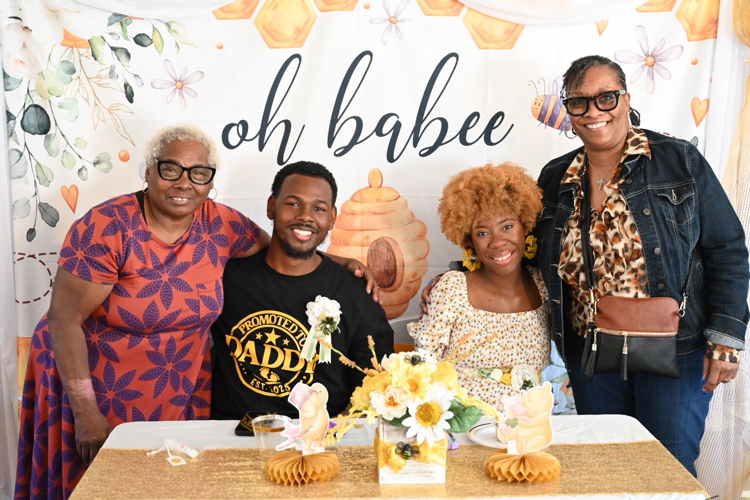 Four guests gather at a bee-themed baby shower with sunflowers and an oh babee decorative banner.