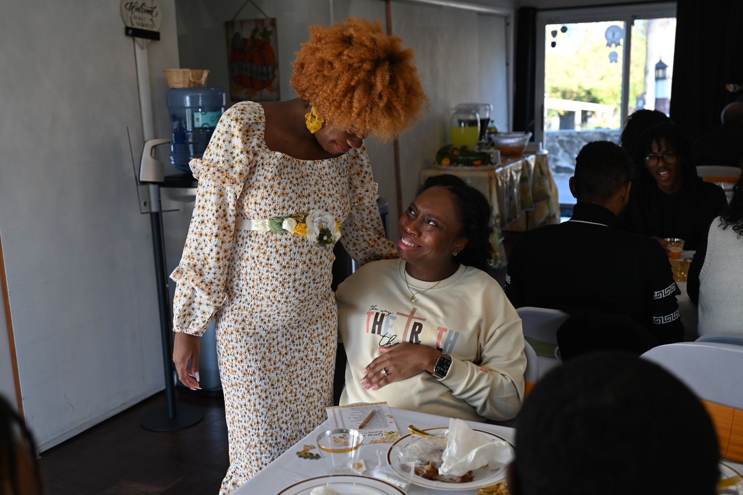 A woman in a floral dress chats with a seated guest at a baby shower event with table settings visible.