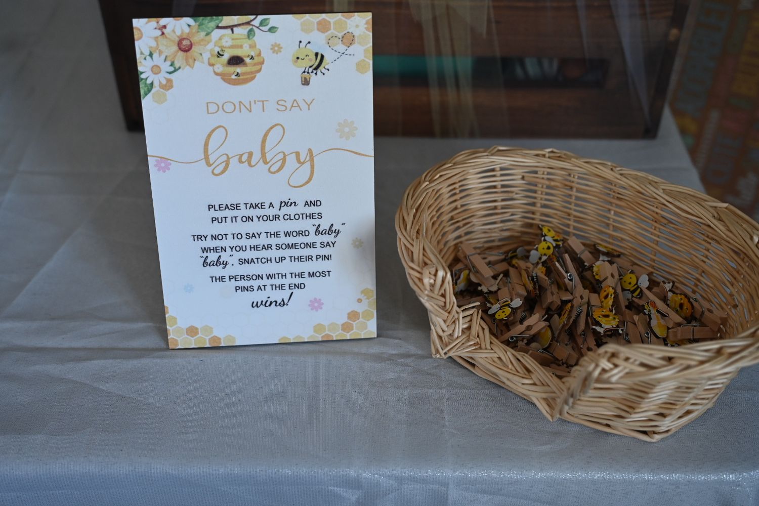 Bee-themed Don't Say Baby game sign displayed next to wicker basket filled with wooden clothespins.