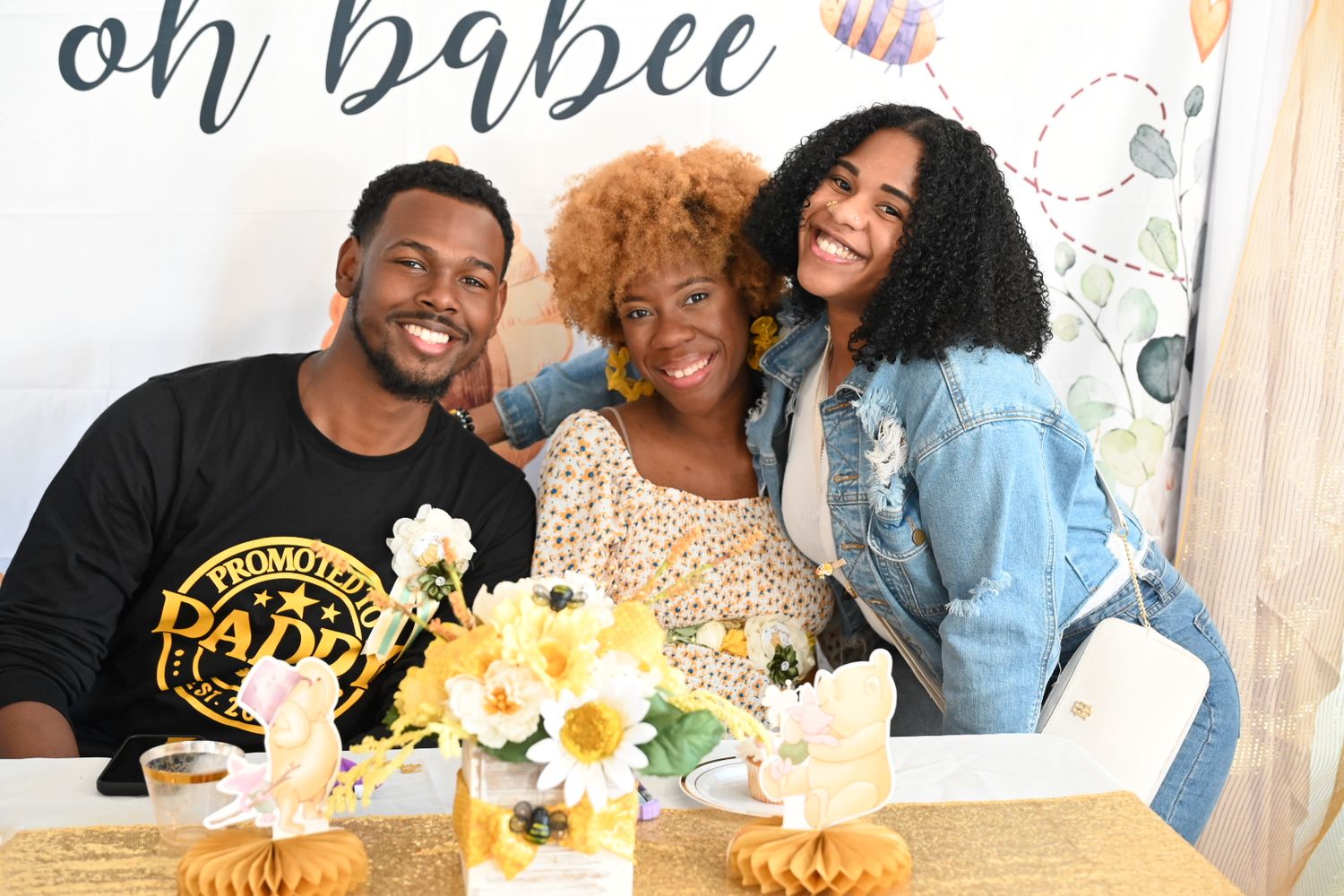 Three guests celebrate at a bee-themed baby shower with sunflowers and an oh babee decorative backdrop.