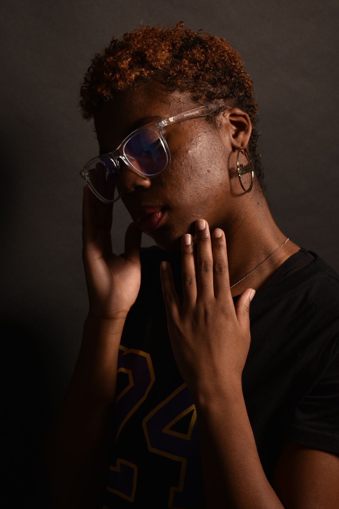 Young woman with short auburn hair holds face thoughtfully wearing clear glasses in moody studio light.