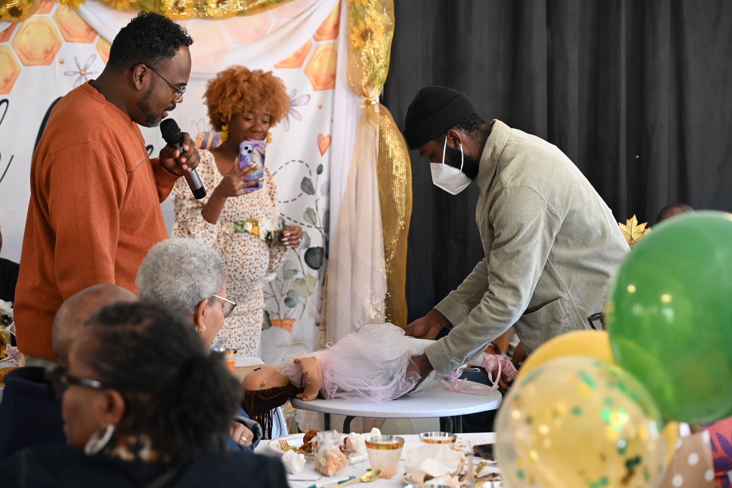 Guests gather around a gift table at a bee-themed baby shower as a man speaks into a microphone.