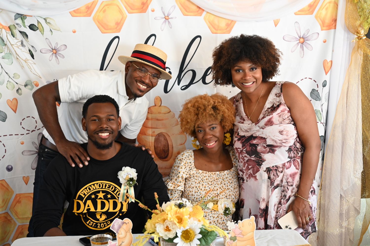 Four people pose together in front of a honeybee-themed baby shower backdrop with flowers.