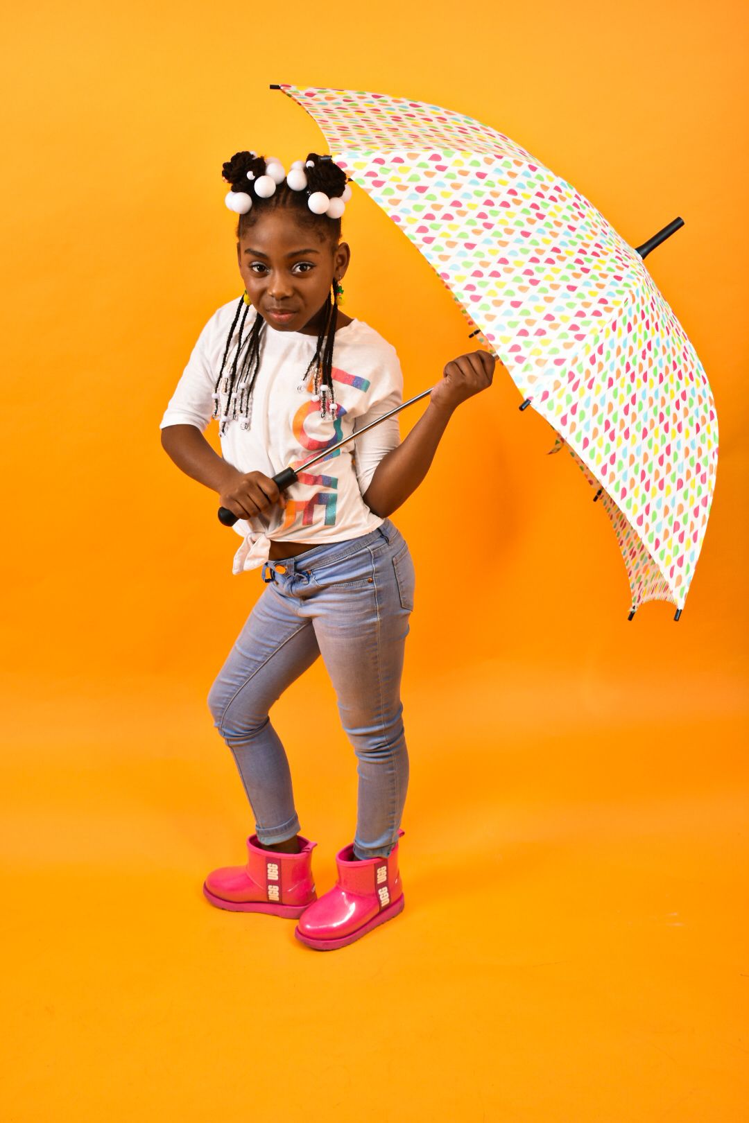 Young Black girl in white shirt and jeans holding colorful umbrella, wearing pink boots on orange background.