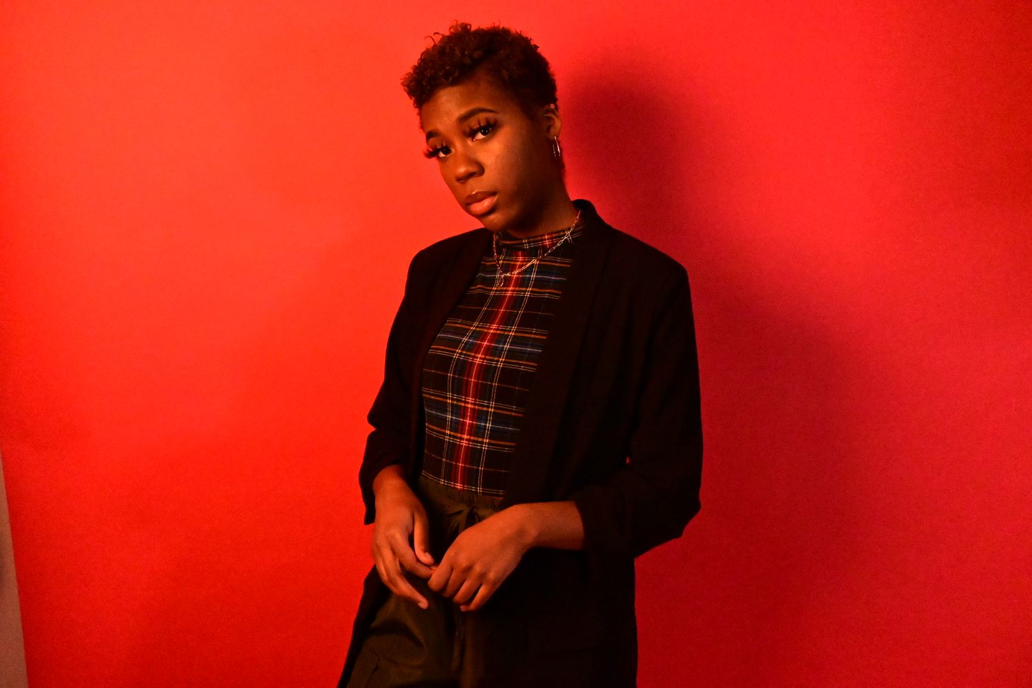 Woman in black blazer and plaid shirt poses against vibrant red background in artistic portrait photography.