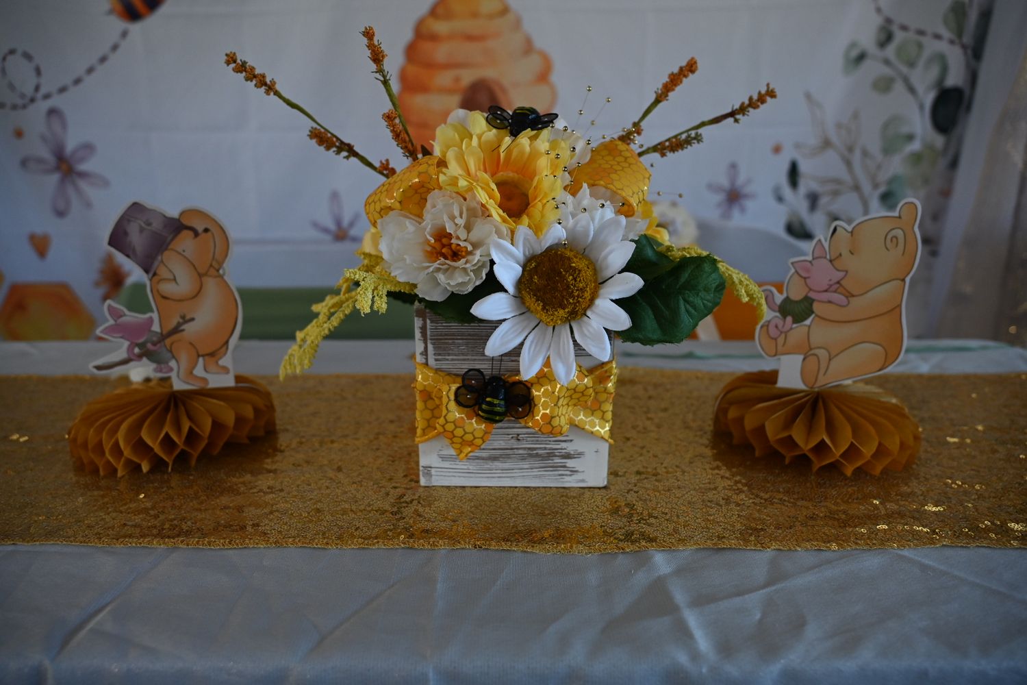 Yellow and white daisy floral centerpiece with honeycomb decorations on gold sequin table runner.
