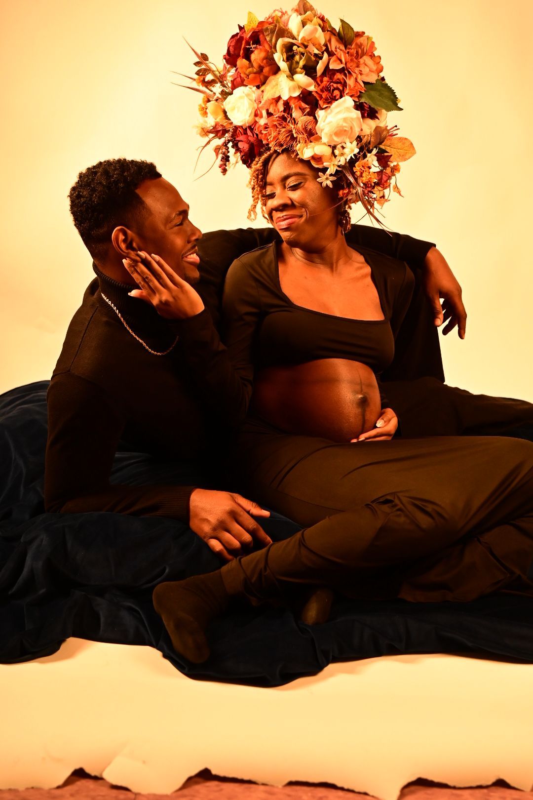 Couple in black outfits posing together with pregnant woman wearing floral crown on warm background.