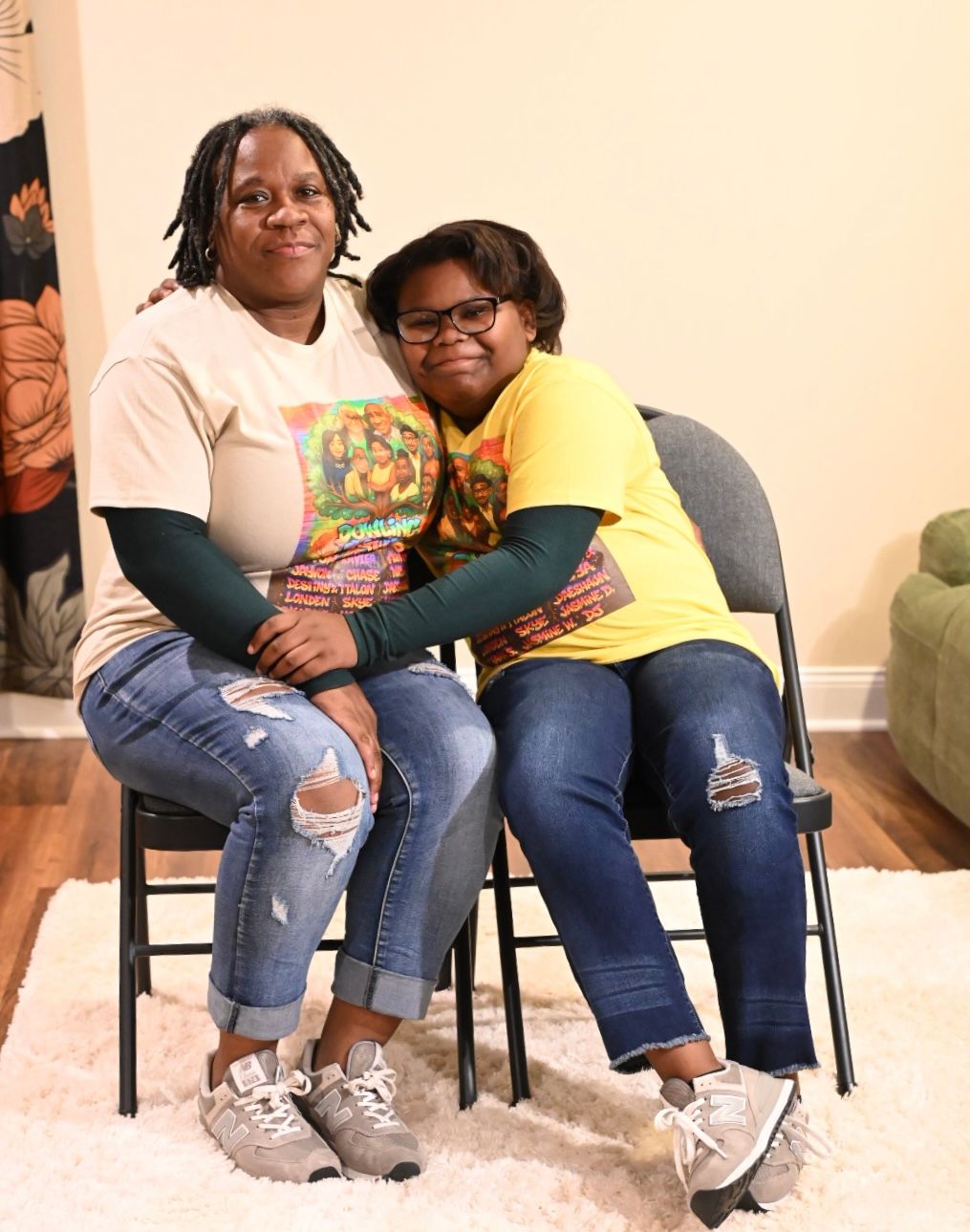 Two women smiling and posing together on chairs, wearing casual colorful t-shirts and jeans in a bright indoor setting.