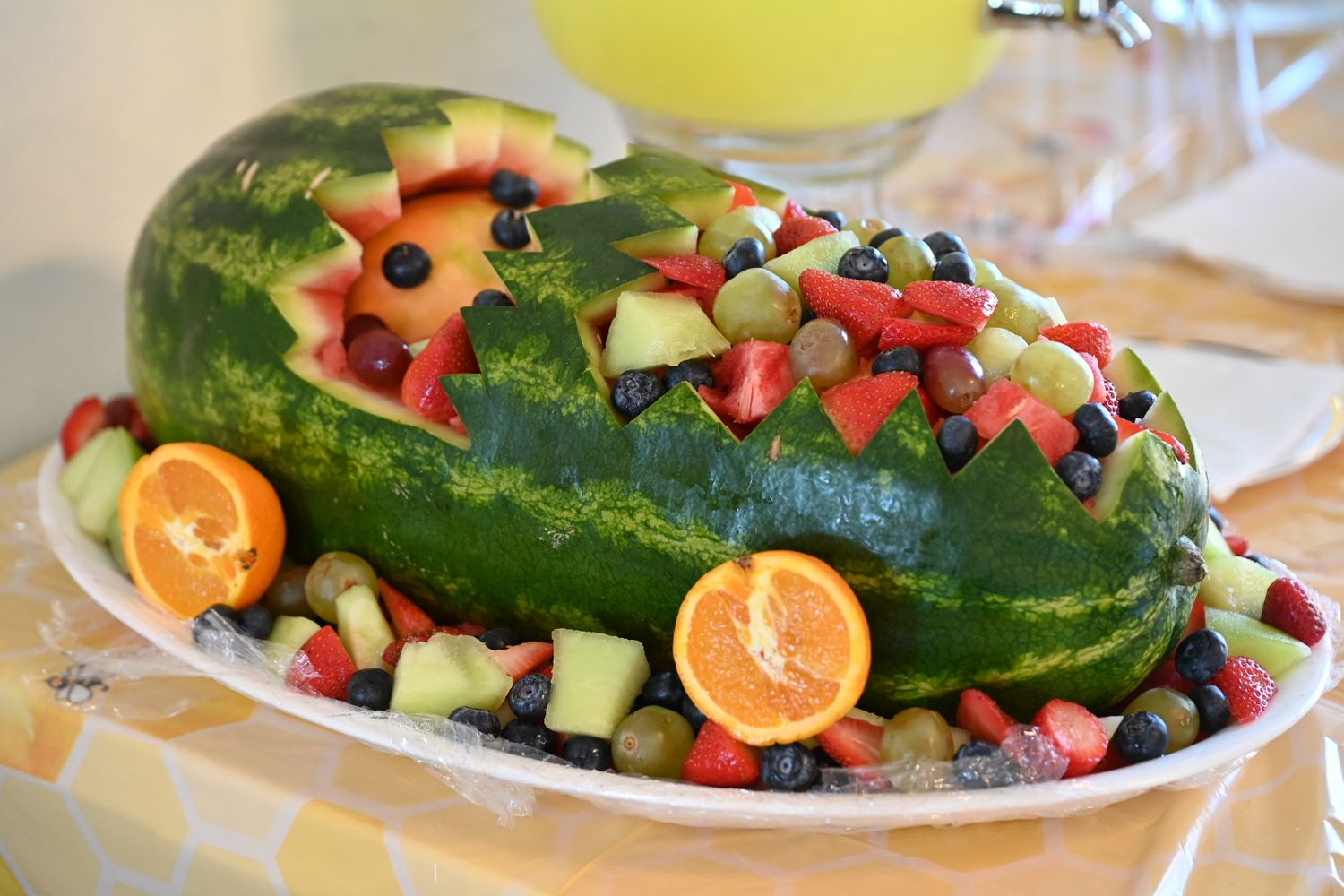 Decorative watermelon carved into a baby carriage shape surrounded by fresh mixed fruit and oranges.