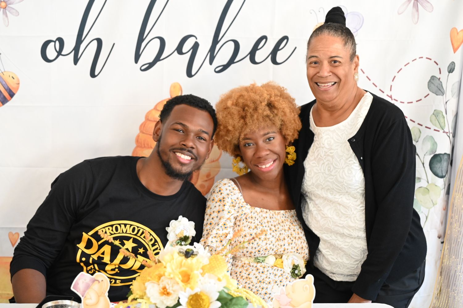 Three people smiling at a baby shower with an oh babee banner and yellow flower arrangements.