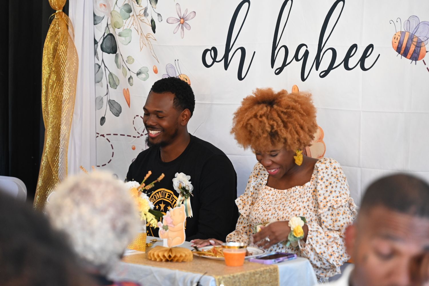A smiling couple sits together at an oh babee bee-themed baby shower celebration.