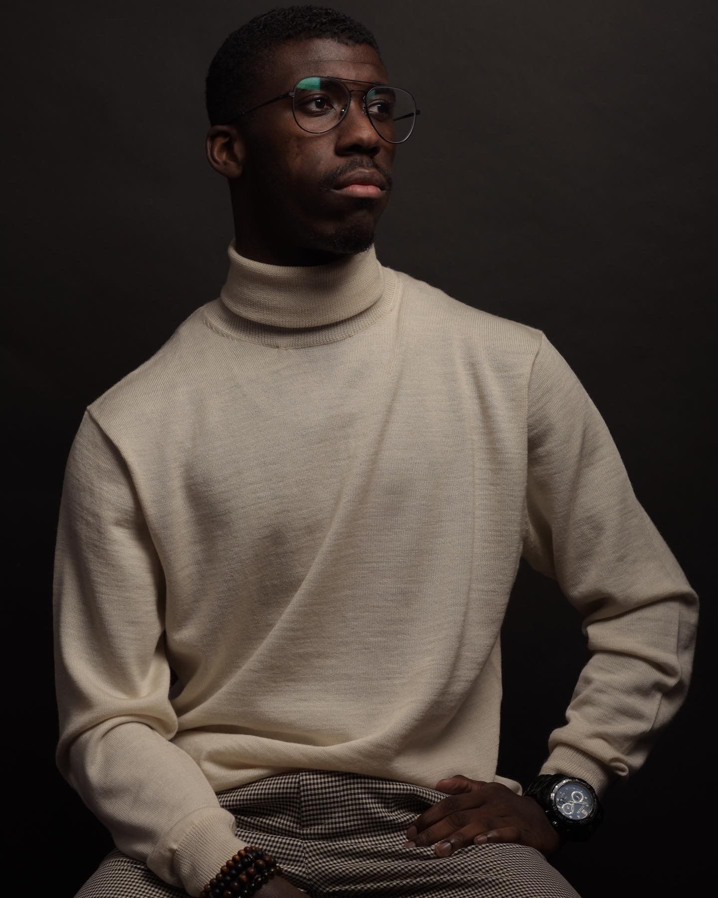 Young Black man in beige turtleneck sweater and glasses sitting confidently against a dark backdrop.
