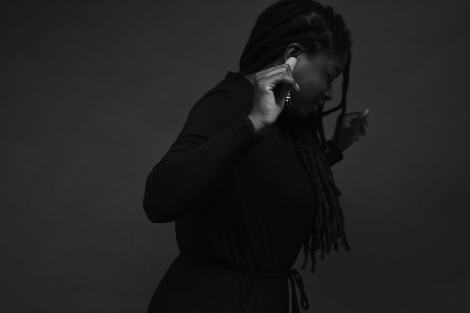 Black and white portrait of a woman with long braids wearing a black outfit, looking down with hand raised to face.