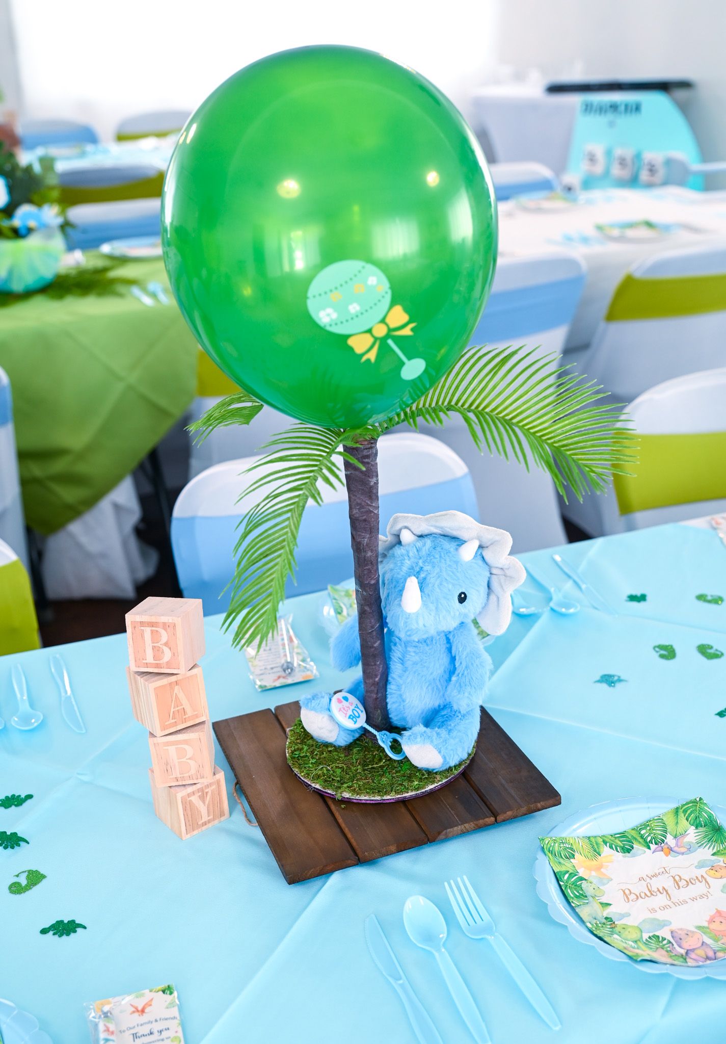 Dinosaur baby shower centerpiece with green balloon, palm tree, blue elephant plush, and letter blocks.