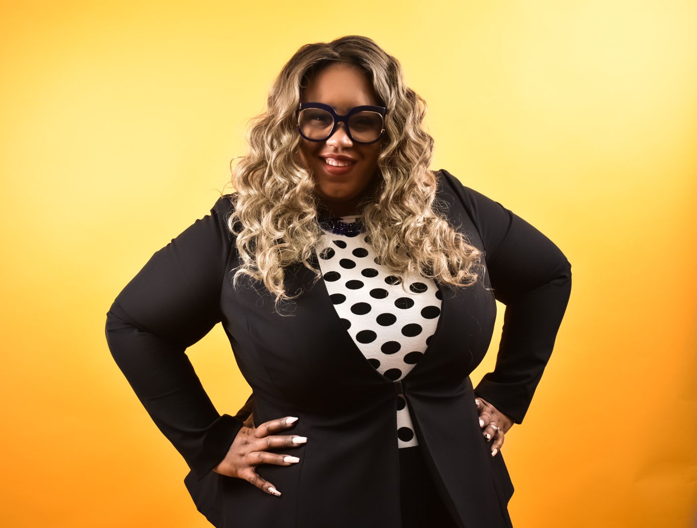 Confident plus-size Black woman in black blazer and polka dot top posing on orange background.