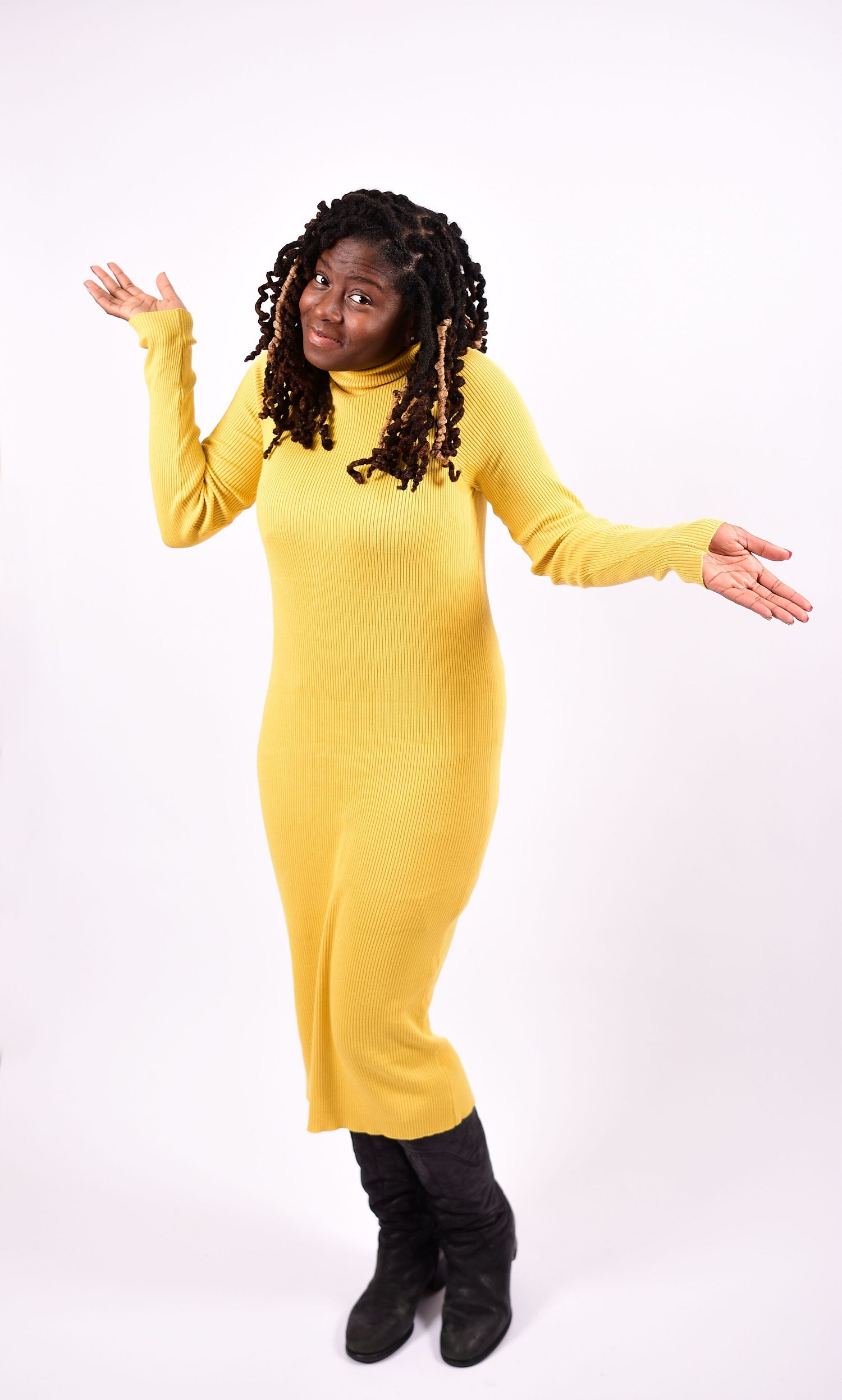 Woman in yellow turtleneck midi dress and black boots posing with open arms.