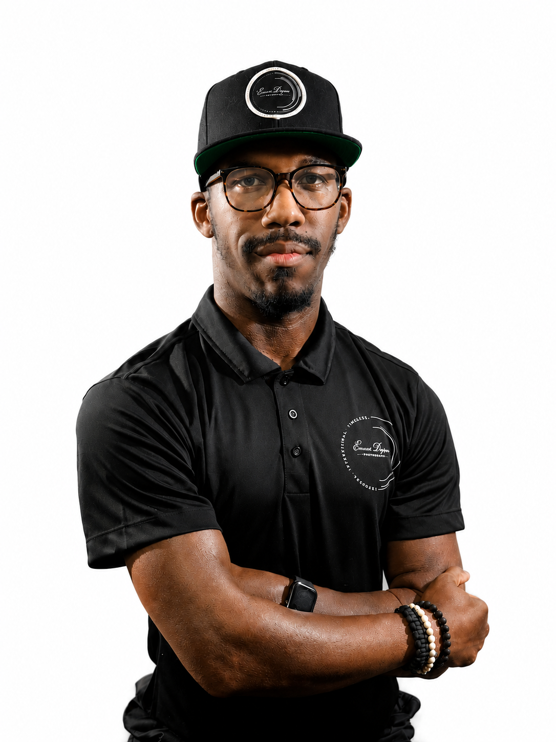 A confident Black man in a black polo shirt and cap stands with arms crossed against a white background.
