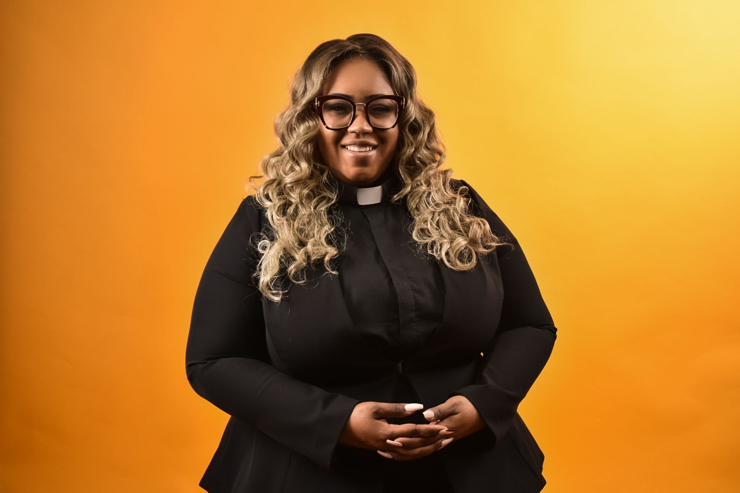 Smiling Black female pastor in black clergy collar shirt and glasses posing against orange background.